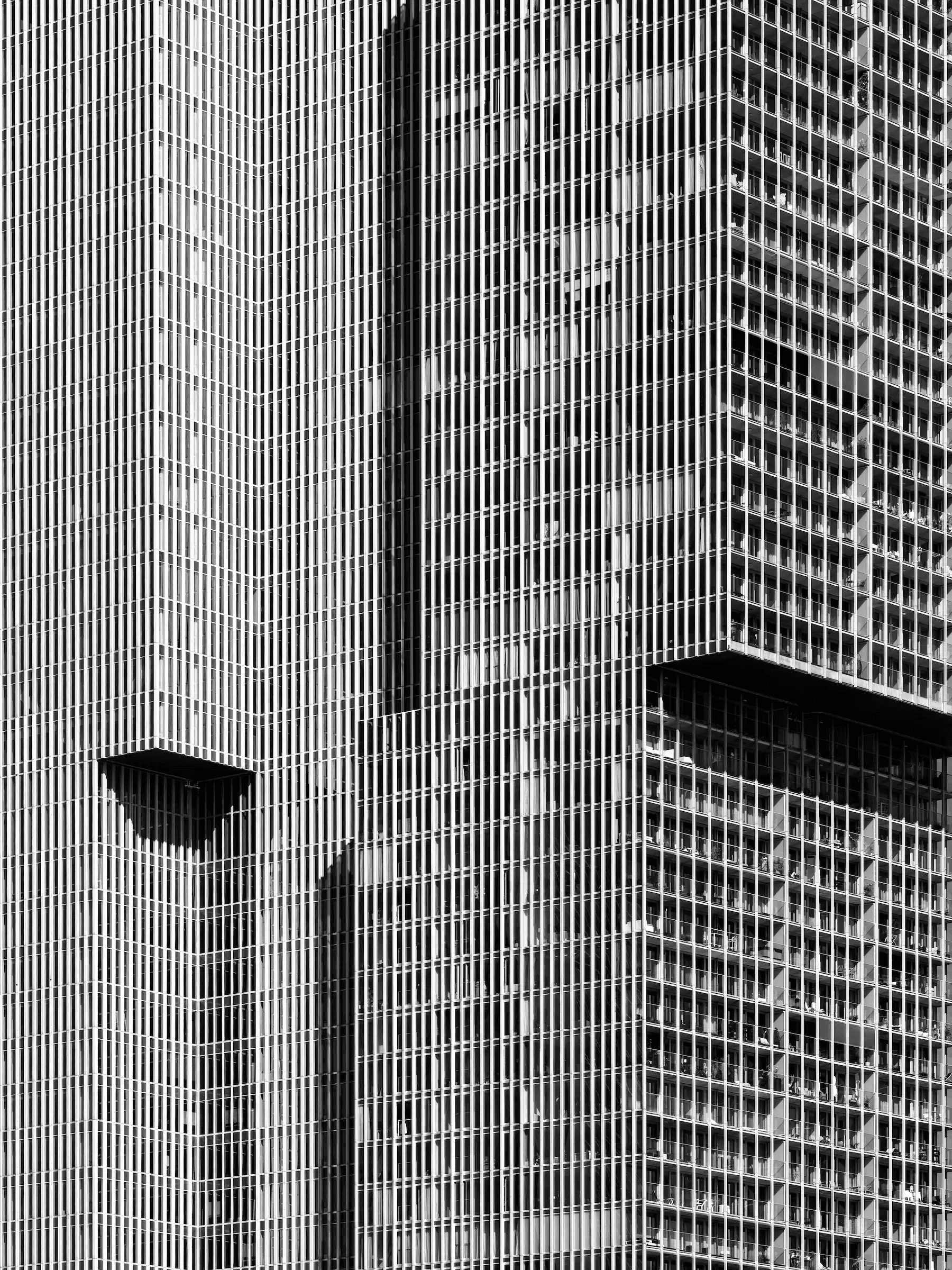 Abstract black and white photograph of the facade of the 'De Rotterdam' building. The image is a tight crop showing the repeating pattern of vertical lines and windows, creating a geometric and textured effect.