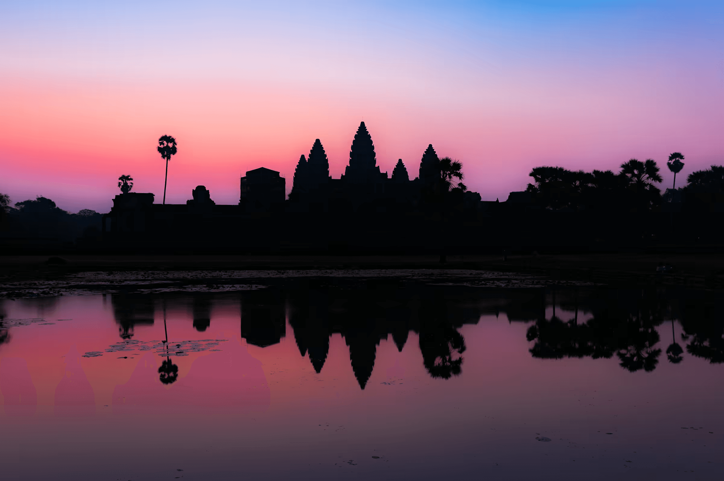 Dawn photograph of Angkor Wat temple complex silhouetted against a gradient sky, with the ancient spires and palm trees perfectly reflected in still water.