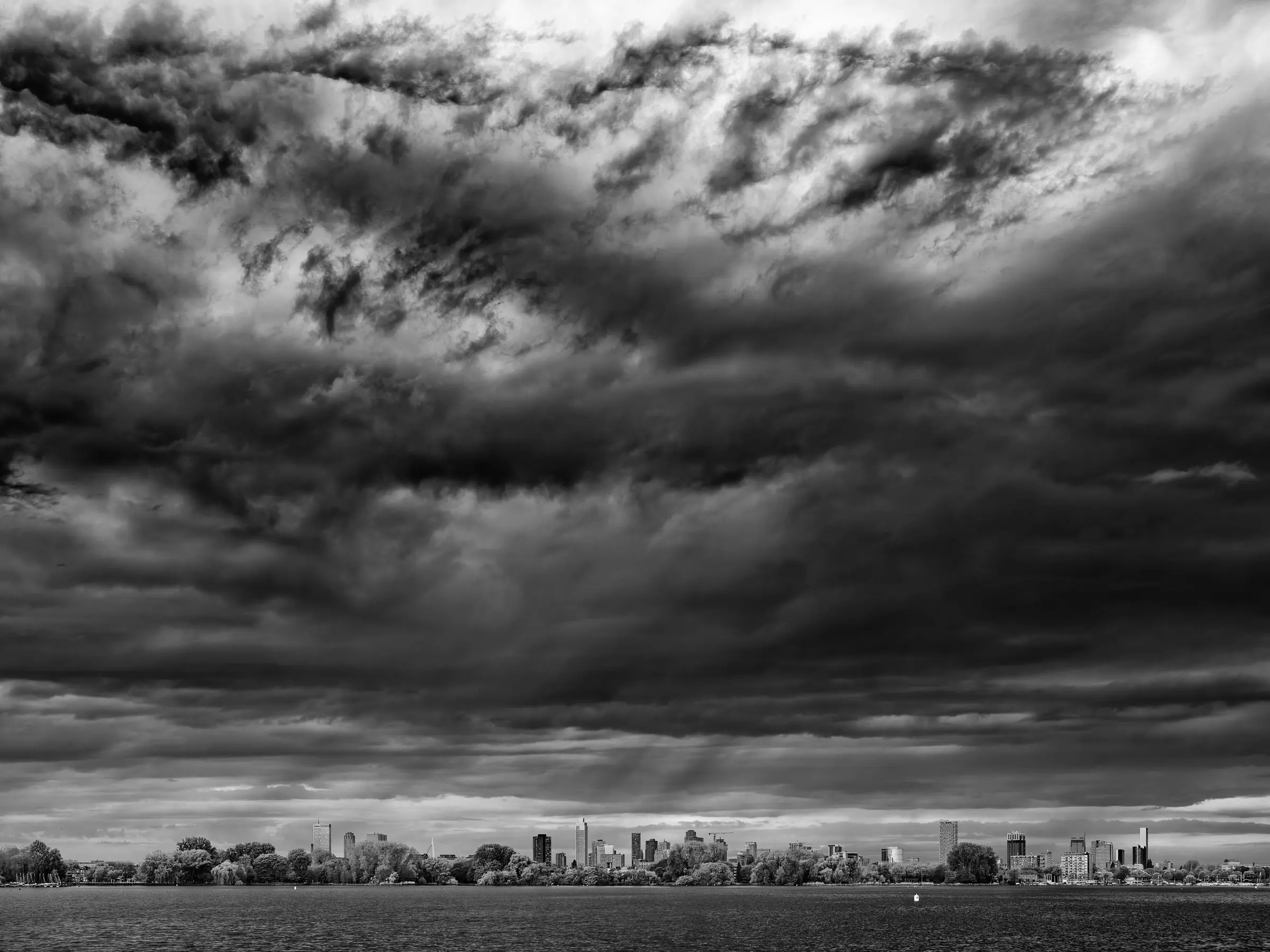 Dramatic black and white photograph of a city skyline beneath heavy, textured storm clouds, seen from across a wide river or lake.