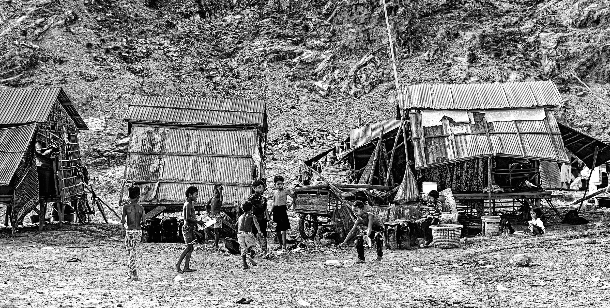 Black and white photograph of several children playing or standing outside simple corrugated metal structures at the base of a rocky hillside.