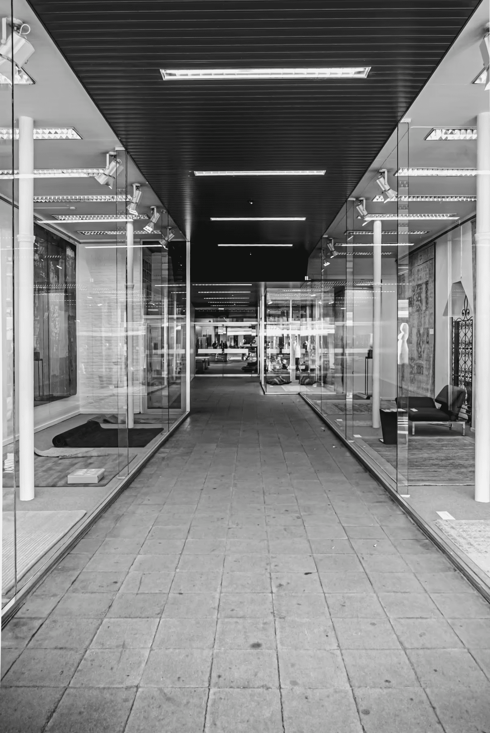 Black and white photograph of a modern city corridor. Strong geometric lines are formed by the architecture, including glass walls and a tiled floor.