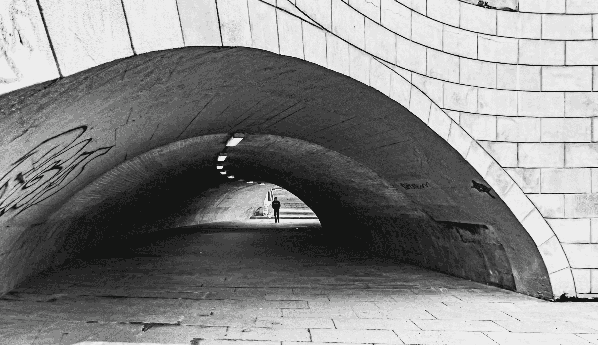 Black and white photograph of an arched city tunnel. The tunnel's brick or stone construction is visible in monochrome.