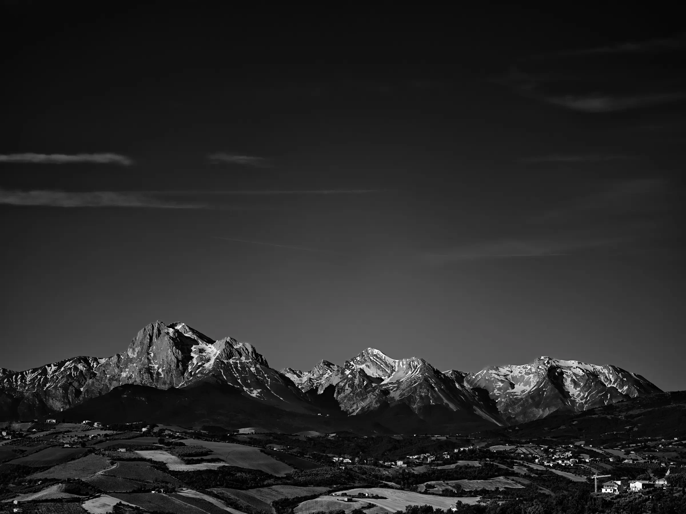 Black-and-white panoramic photograph of snow-covered mountains above a rural valley under a large dark sky with thin clouds.