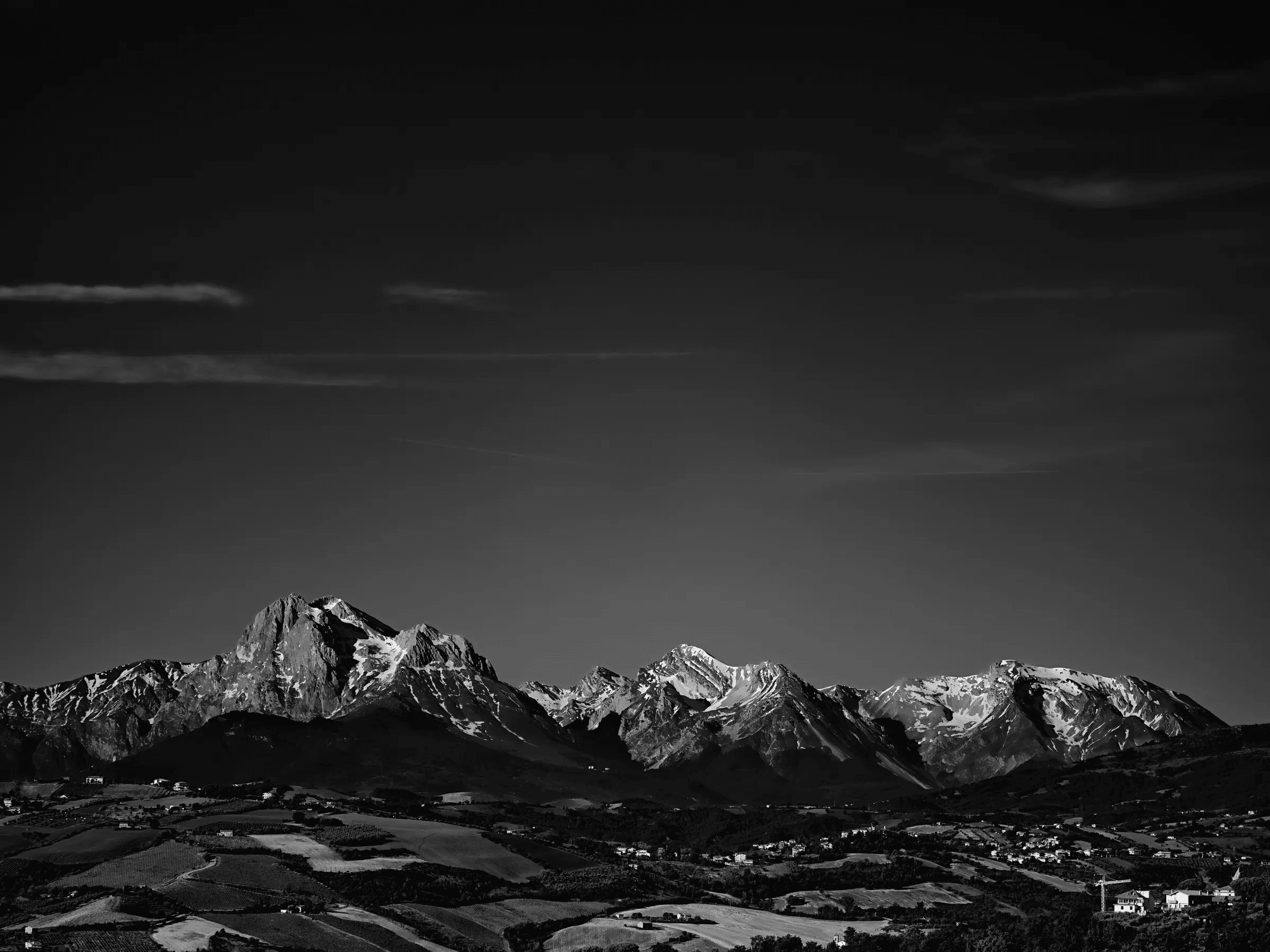 Black-and-white panoramic photograph of snow-covered mountains above a rural valley under a large dark sky with thin clouds.