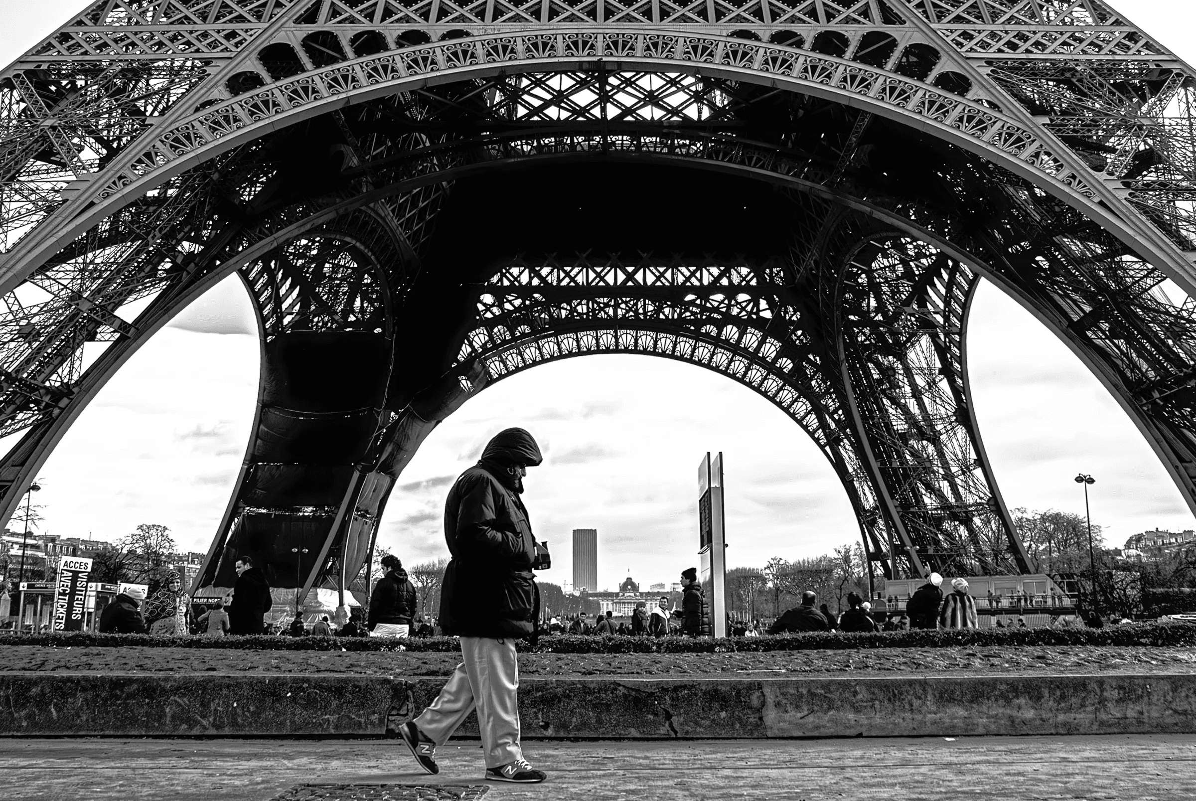 Black and white photograph of the Eiffel Tower in Paris, France, taken from a low angle.  A person is walking in the foreground, providing scale.