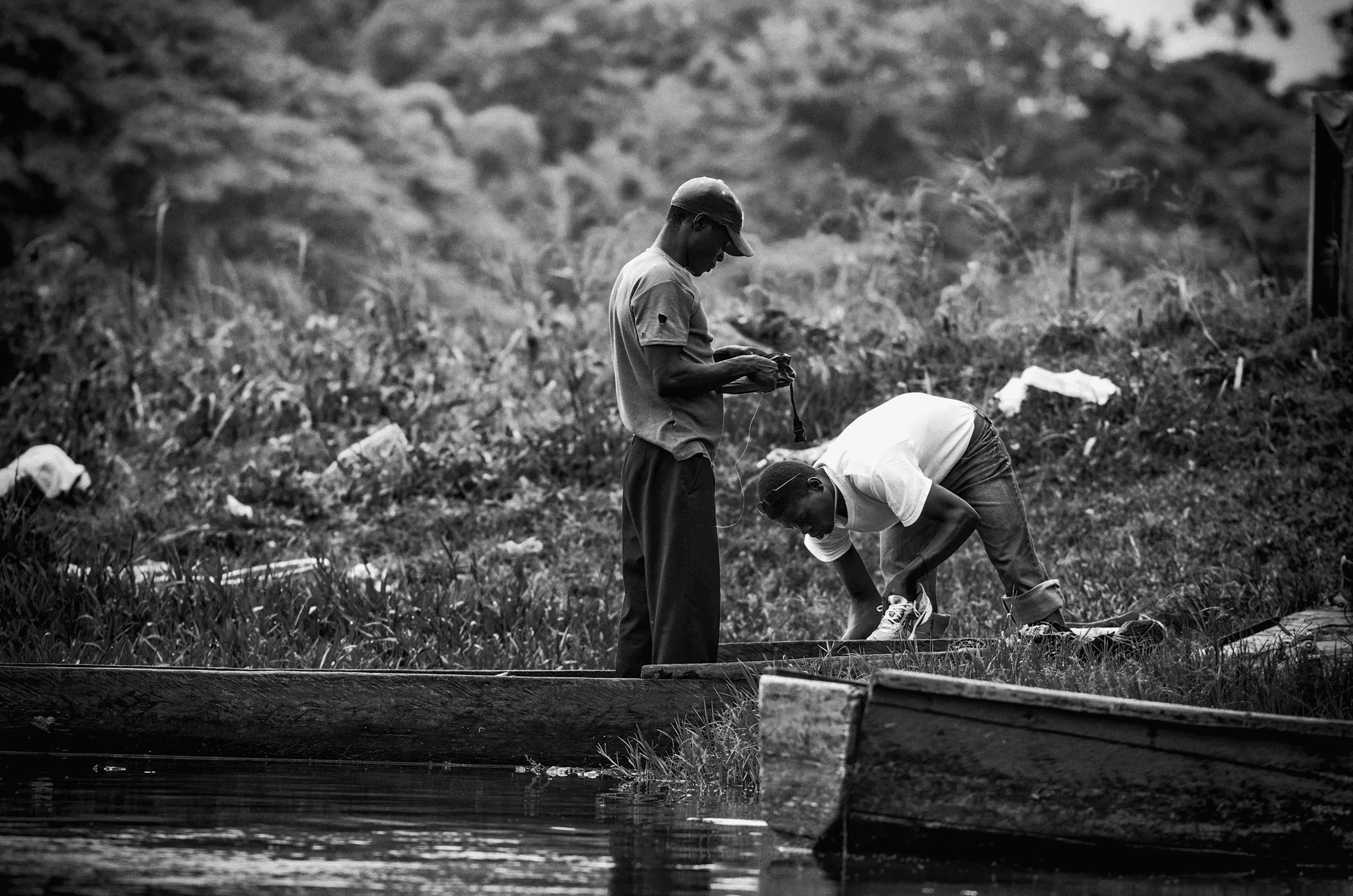 Click to view in fullscreen Two men working on or near simple boats/canoes on a riverbank, possibly preparing fishing gear.
