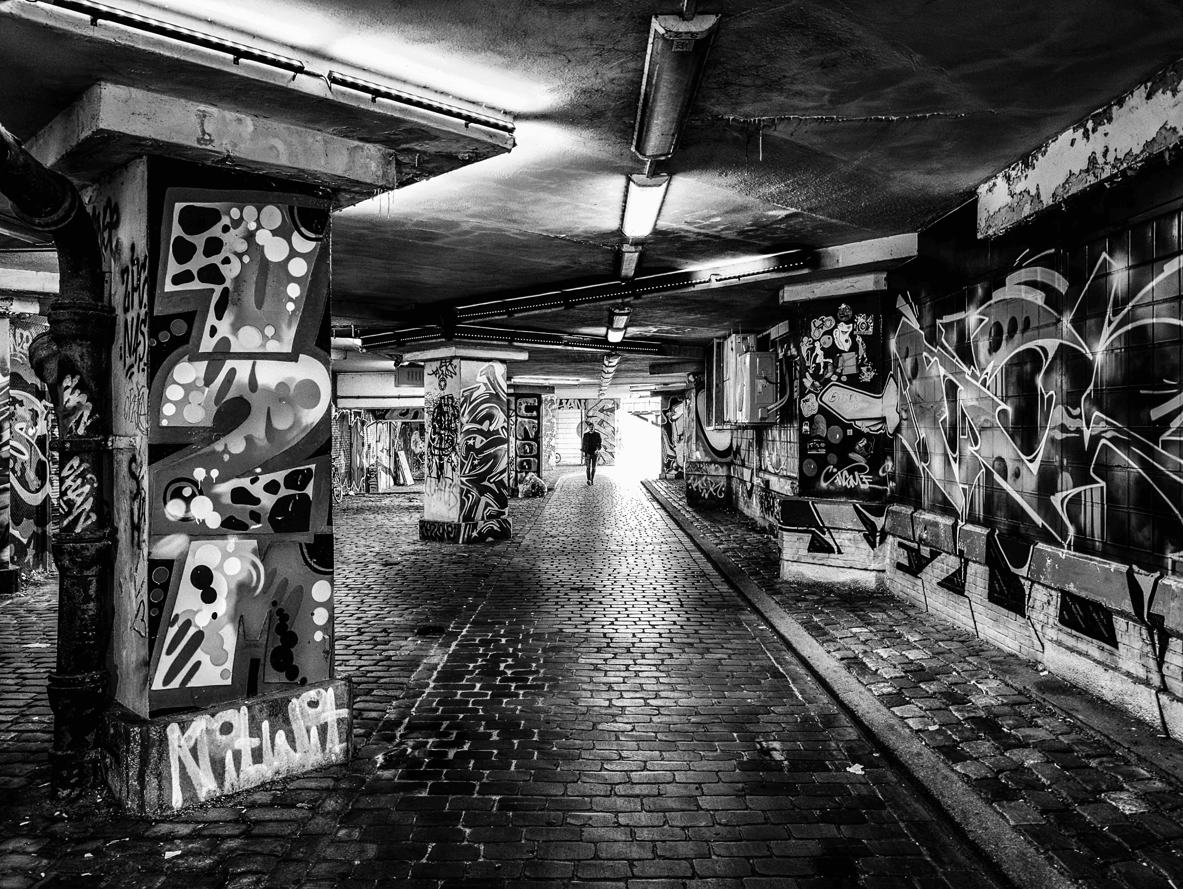 Black and white photograph of a person walking through a graffiti-covered urban underpass with strong overhead lighting and wet cobblestones.
