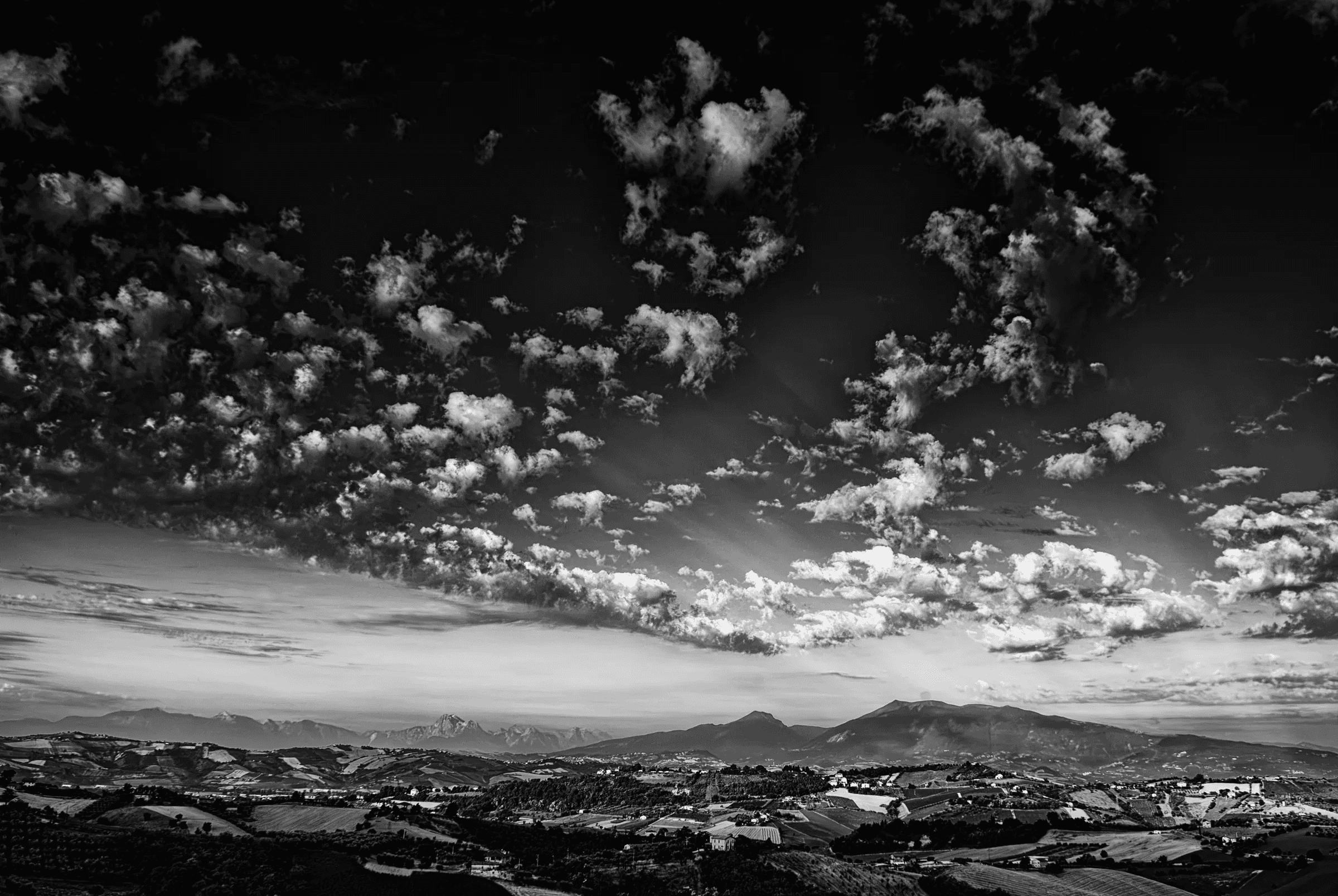 Expansive black and white landscape photograph showing rolling hills of the Marche region with the Gran Sasso mountain range visible in the distance under a dramatic sky filled with scattered clouds..
