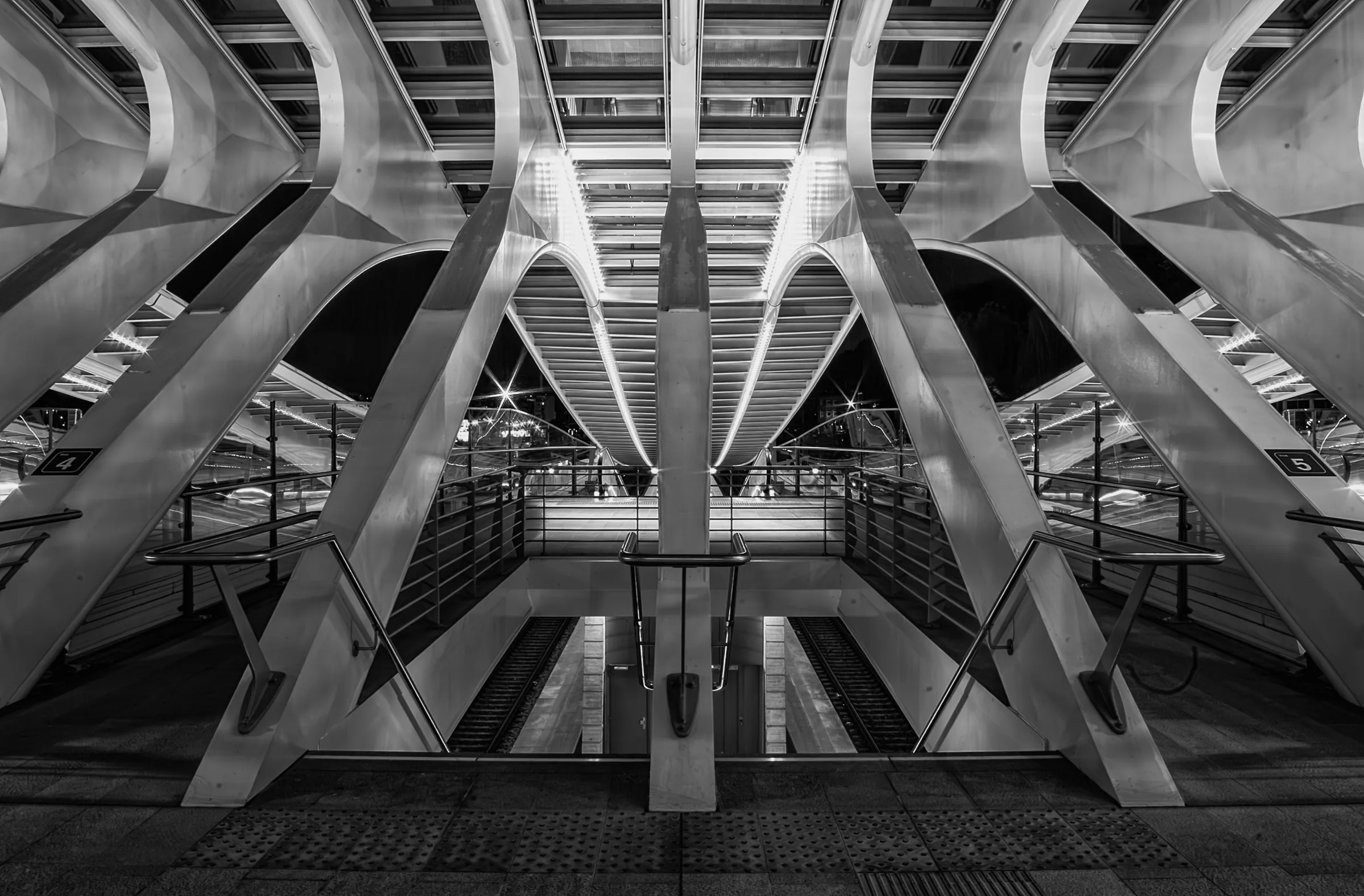 Symmetrical black and white photograph of the interior of Liège-Guillemins railway station, designed by Santiago Calatrava, showing the arched roof supports, platforms, and walkways.