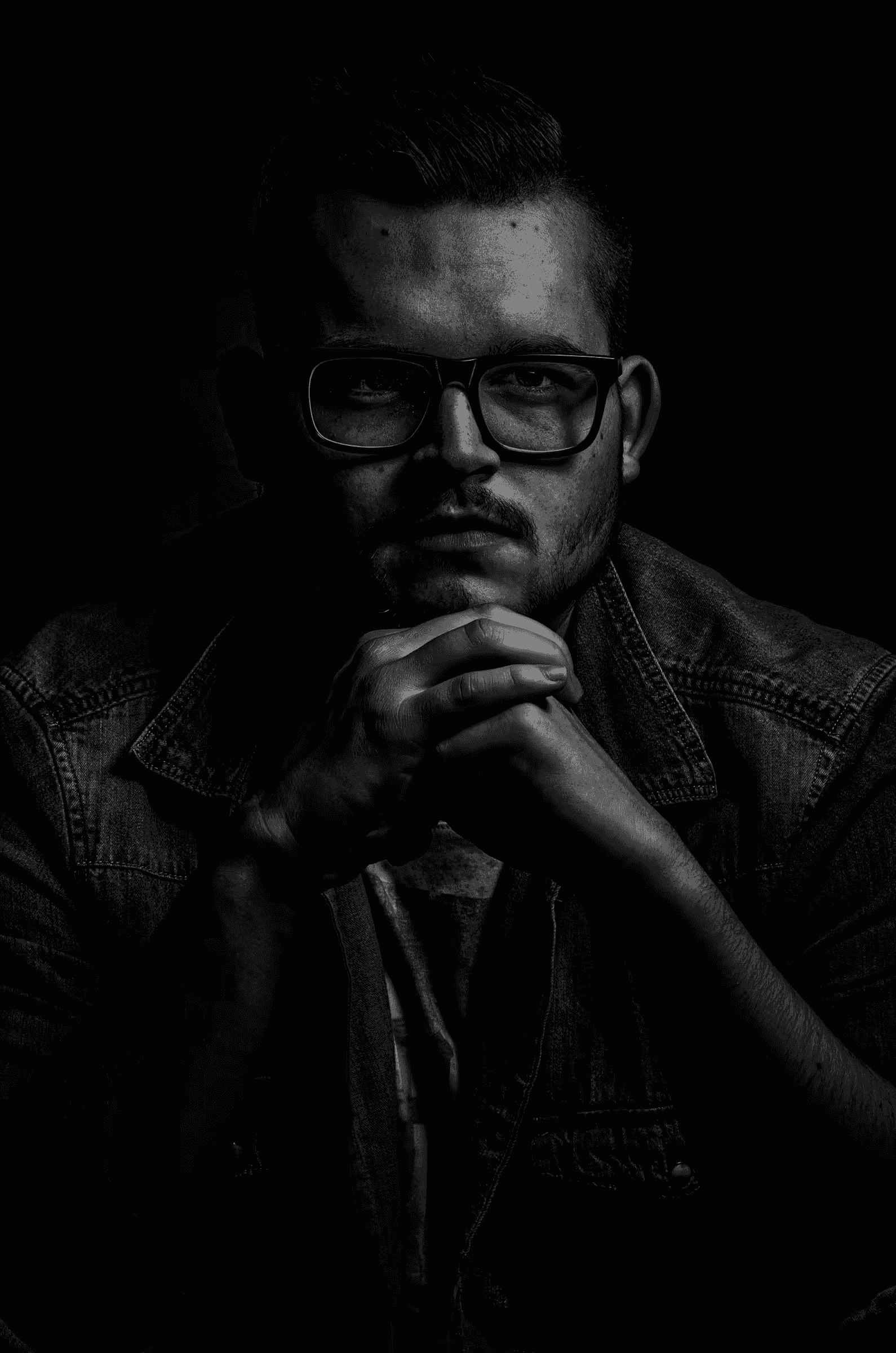 Low-key black and white portrait photograph of a man wearing glasses. The subject is positioned to the right side of the frame, his face partially illuminated, with the rest of the image in deep shadow.