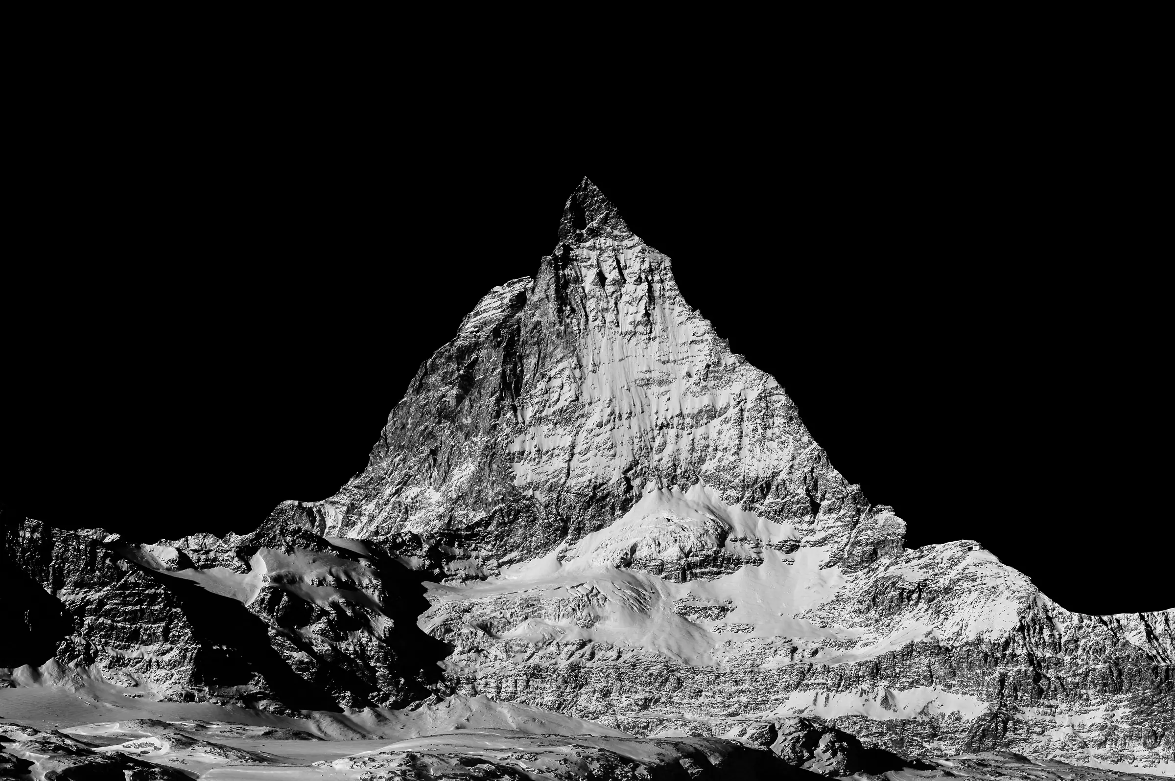 Black-and-white photograph of the Matterhorn mountain with sharp contrast against a deep black sky, highlighting the rugged textures and snow-covered slopes.