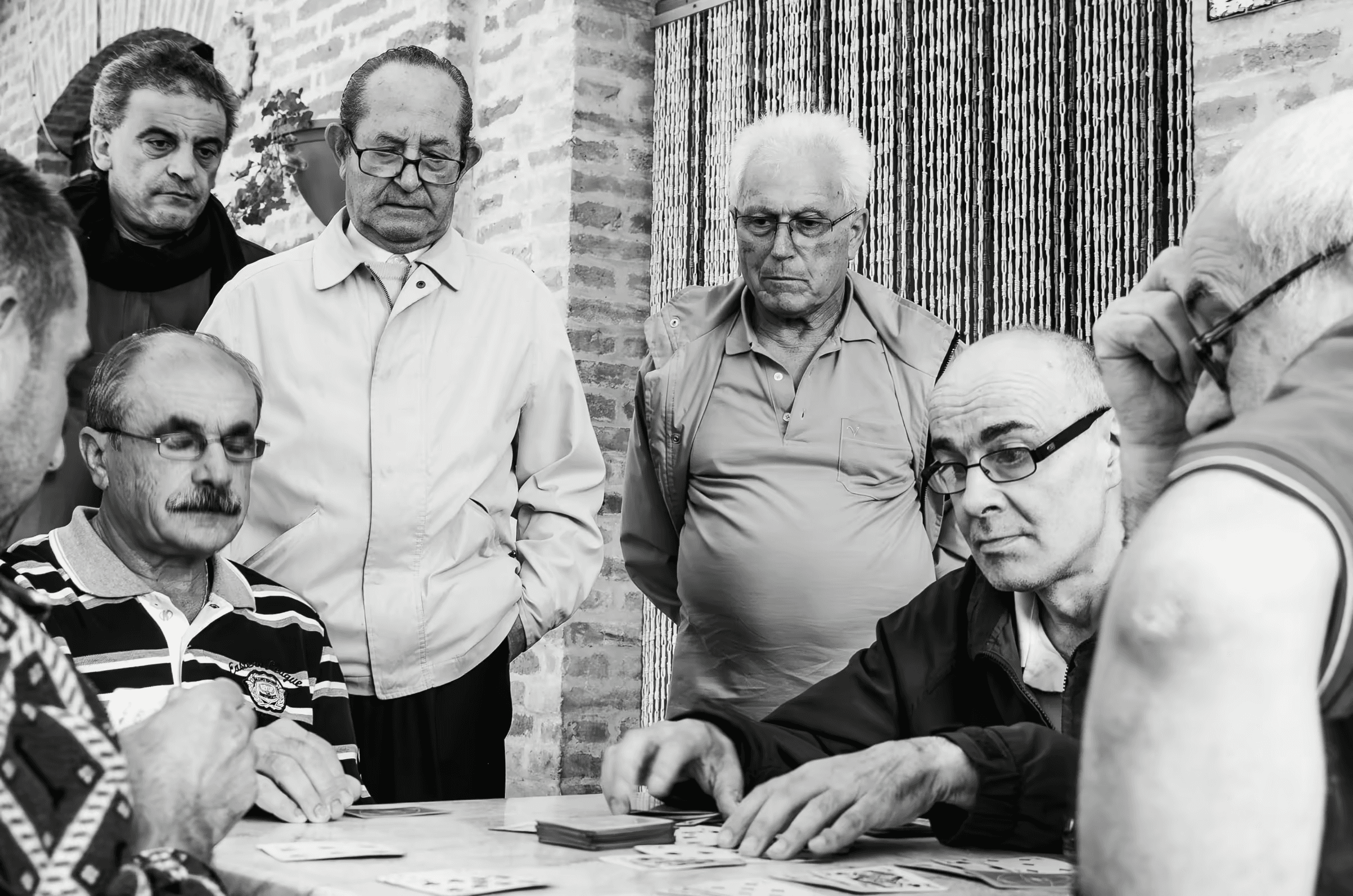 Black and white photograph of a group of older men playing cards. The men are intently focused on their cards and the game, their expressions serious and engaged.
