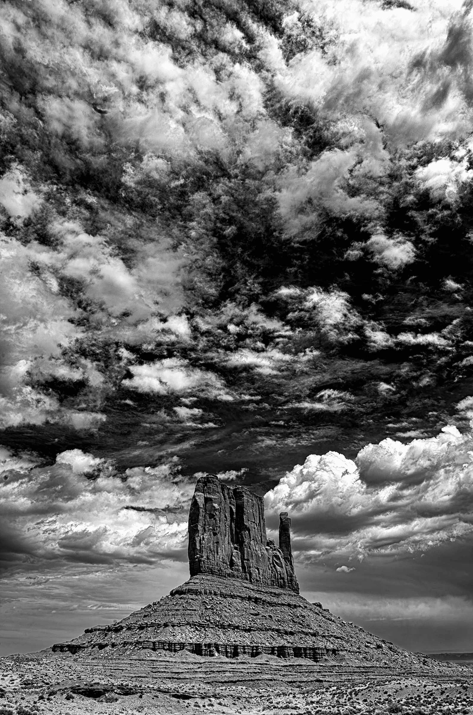 Click to view in fullscreen Black and white photograph of a solitary sandstone butte in Monument Valley beneath a dramatic, textured sky filled with layered clouds.
