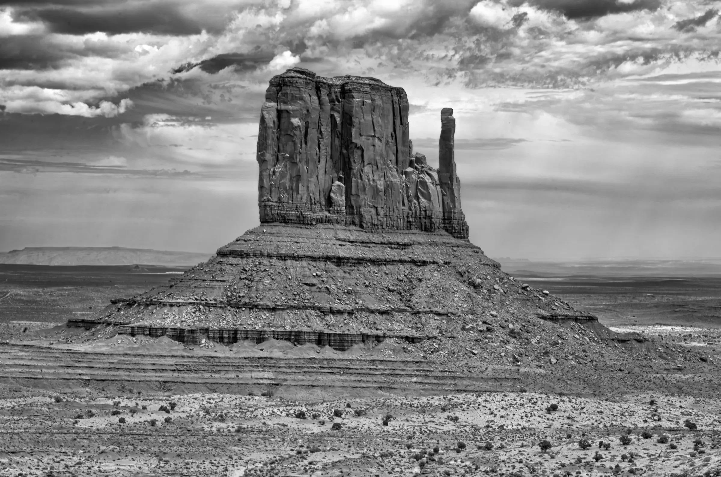 A massive sandstone butte stands alone in a wide desert plain under dramatic, cloud-filled skies. The image is in black and white.