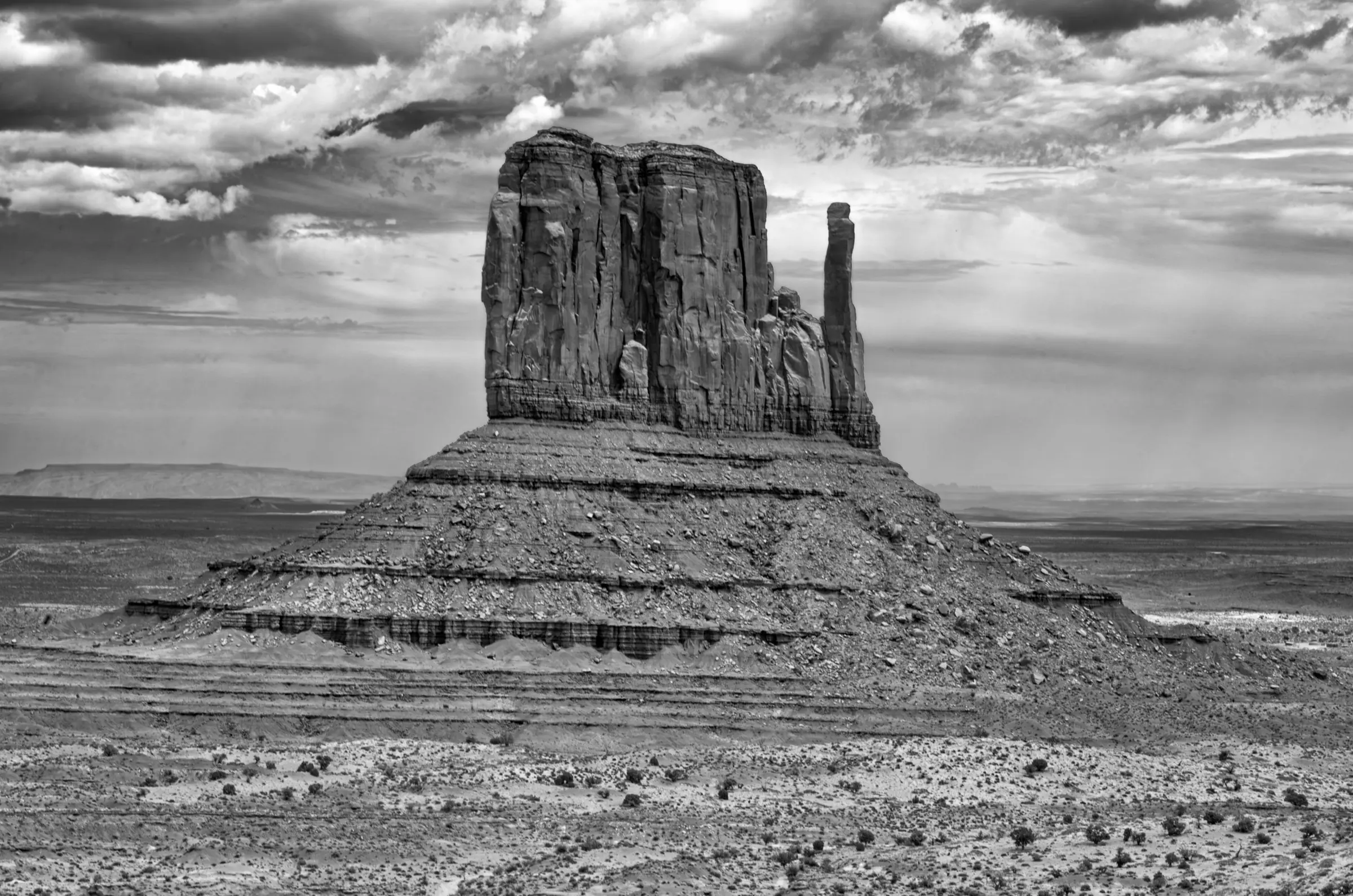 A massive sandstone butte stands alone in a wide desert plain under dramatic, cloud-filled skies. The image is in black and white.