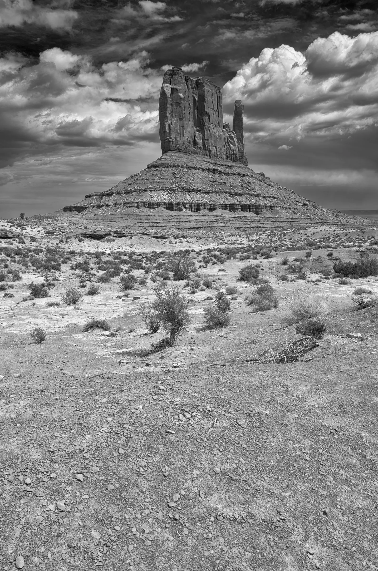 A black and white photograph of a large sandstone butte in Monument Valley, with dramatic clouds overhead and sparse desert vegetation in the foreground.