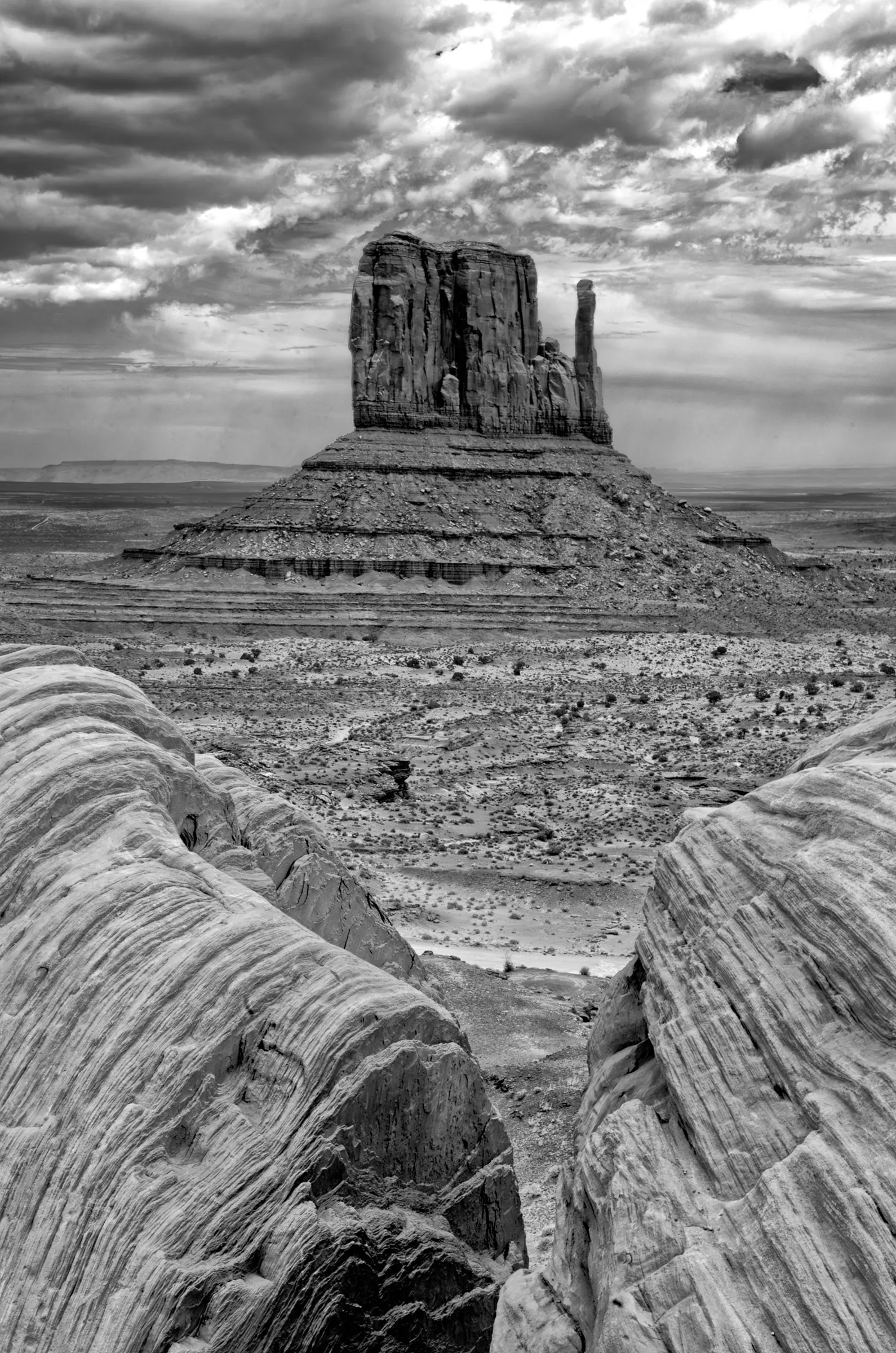 Black and white photograph of a large sandstone butte in a desert landscape, framed by textured foreground rocks and dramatic, cloud-filled sky.