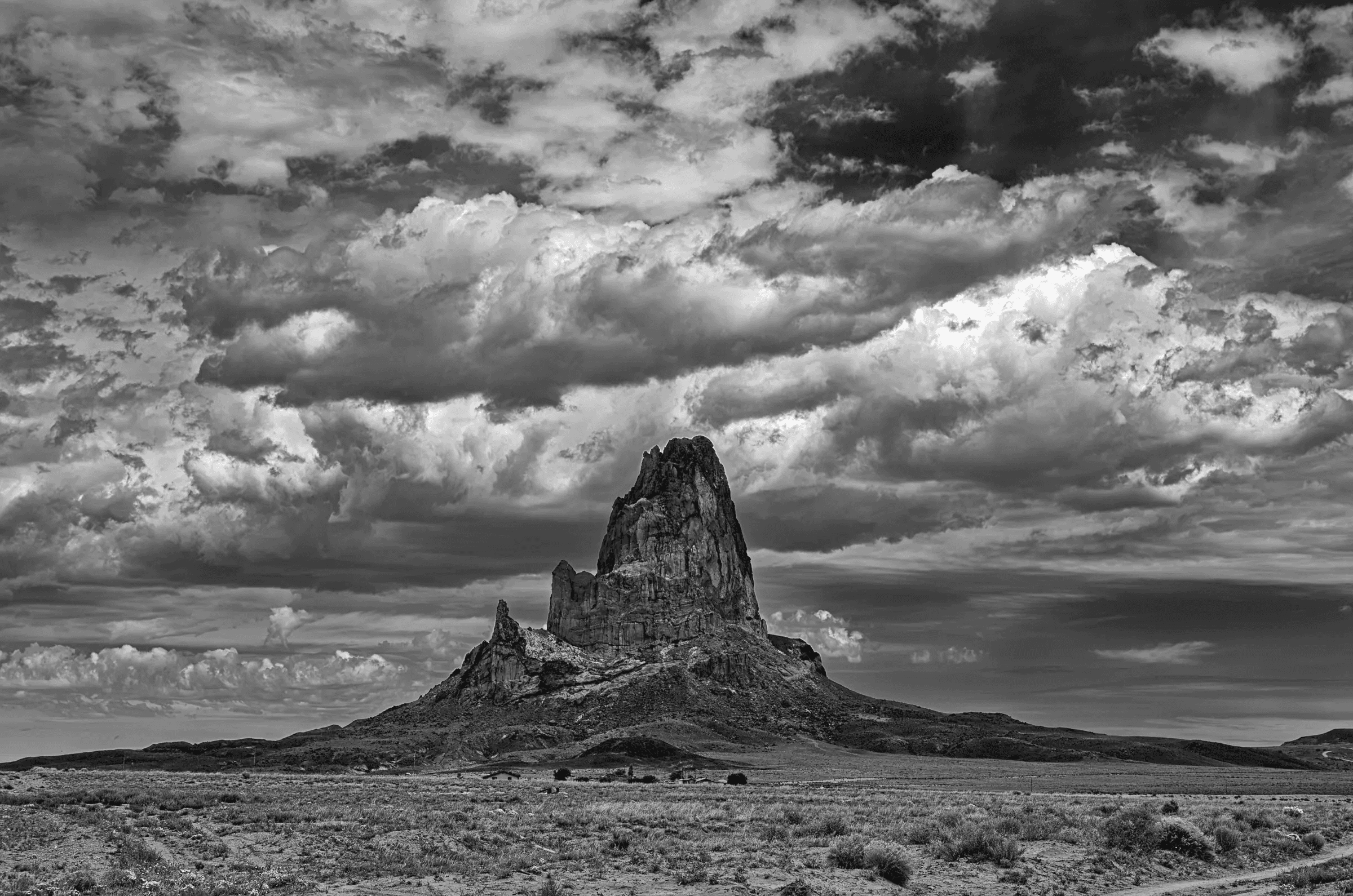 Click to view in fullscreen A dramatic black and white photograph of a towering rock formation rising from arid plains beneath a vast, textured sky filled with heavy, swirling clouds.