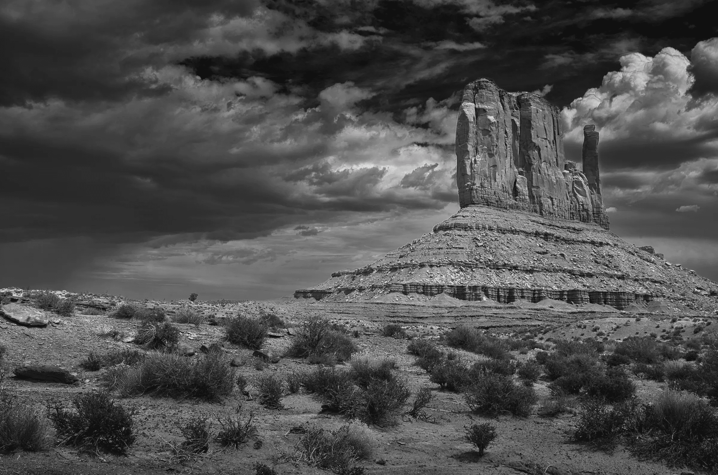 Black and white photograph of a massive desert butte beneath dramatic, textured storm clouds, with sparse scrub in the foreground.