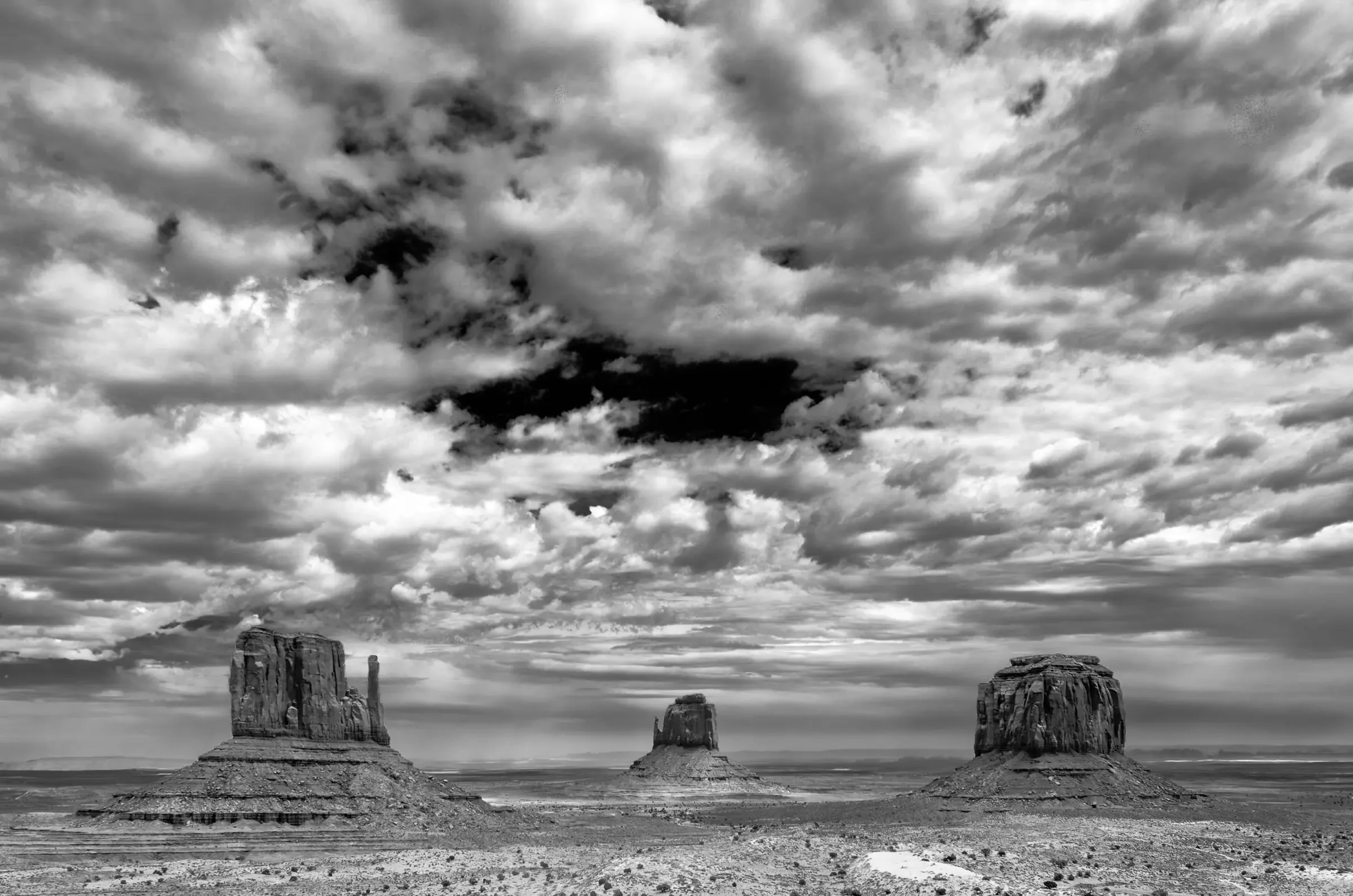 Three large rock formations rise from a flat desert plain beneath a dramatic, cloud-filled sky in black and white.