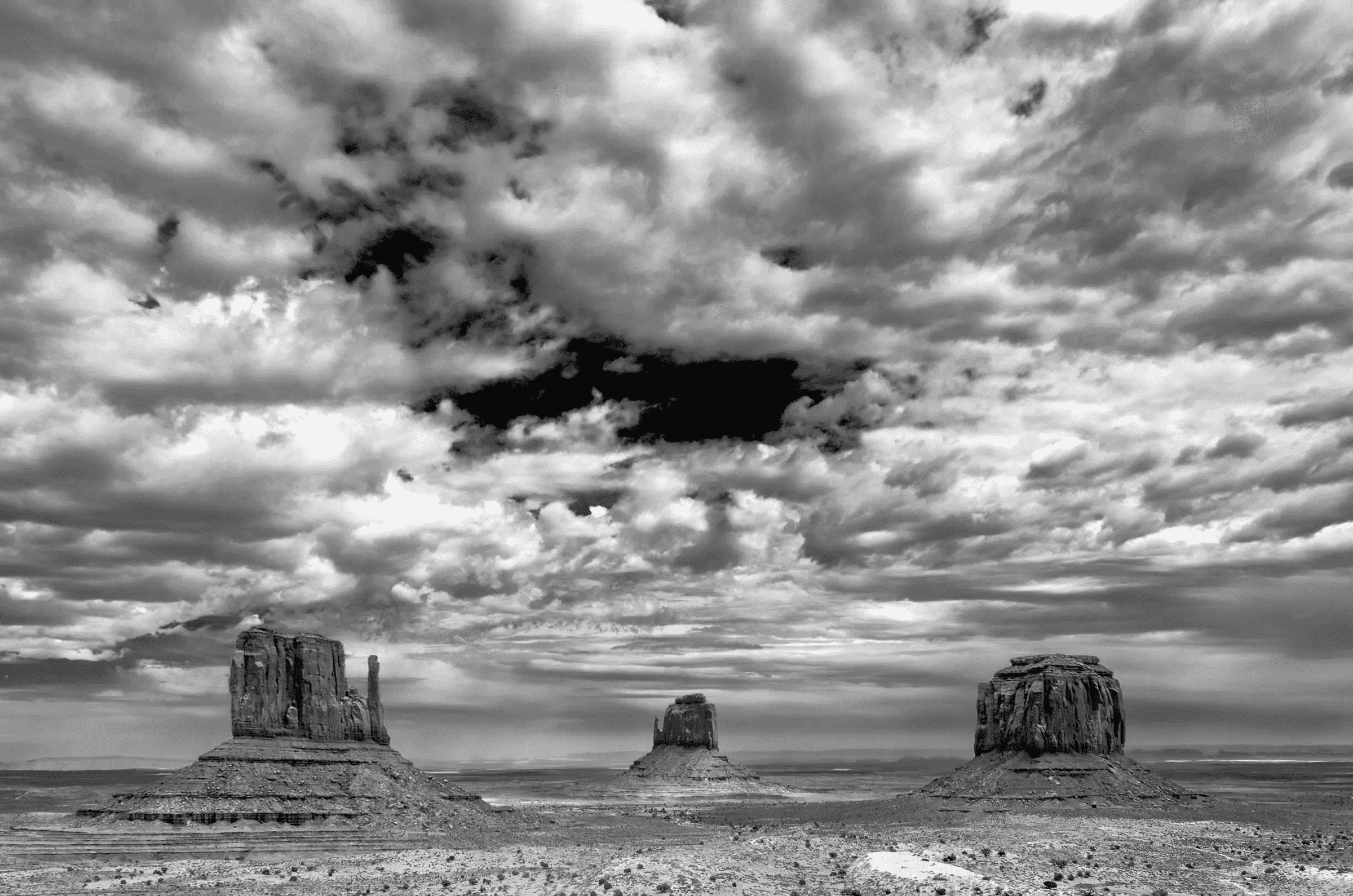 Monumental Weather Three large rock formations rise from a flat desert plain beneath a dramatic, cloud-filled sky in black and white.