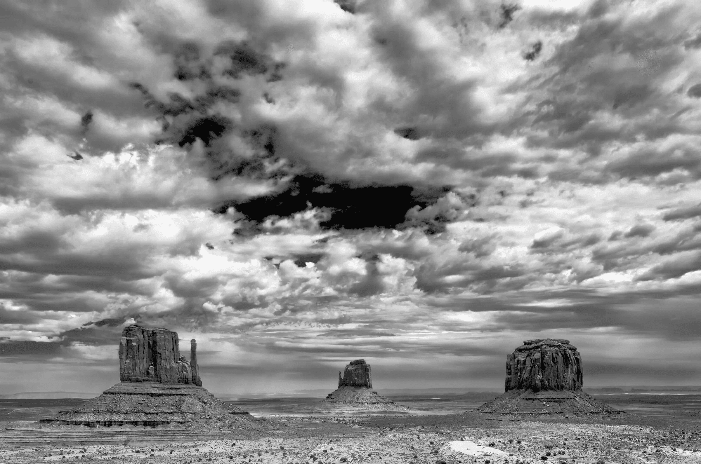 Click to view in fullscreen Three large rock formations rise from a flat desert plain beneath a dramatic, cloud-filled sky in black and white.