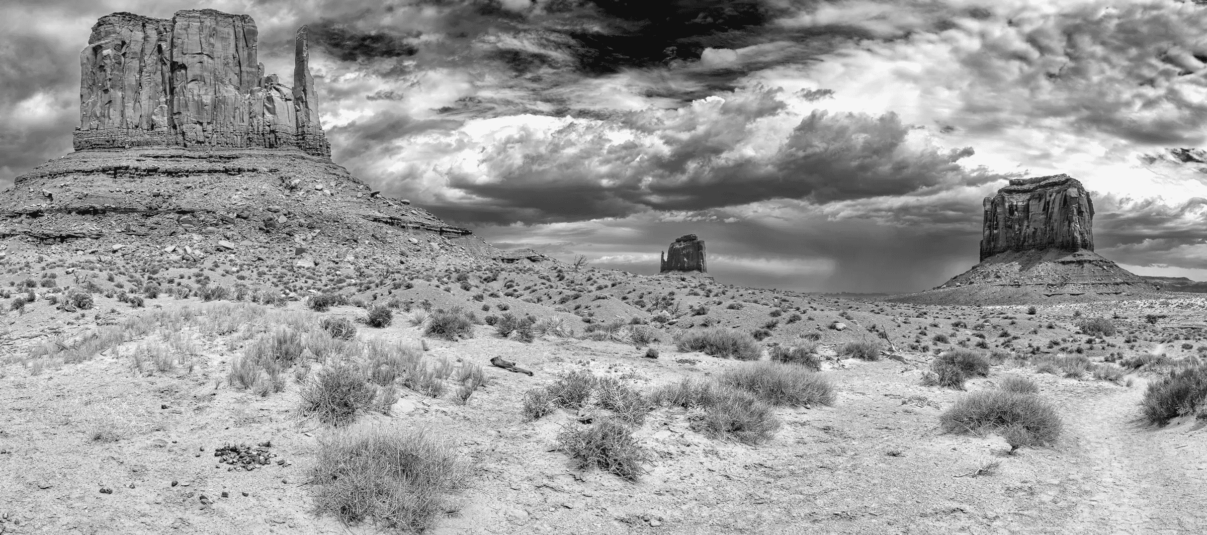 Monuments Under Weather Black and white panorama of Monument Valley with three large buttes rising from arid desert, beneath dramatic, textured clouds.
