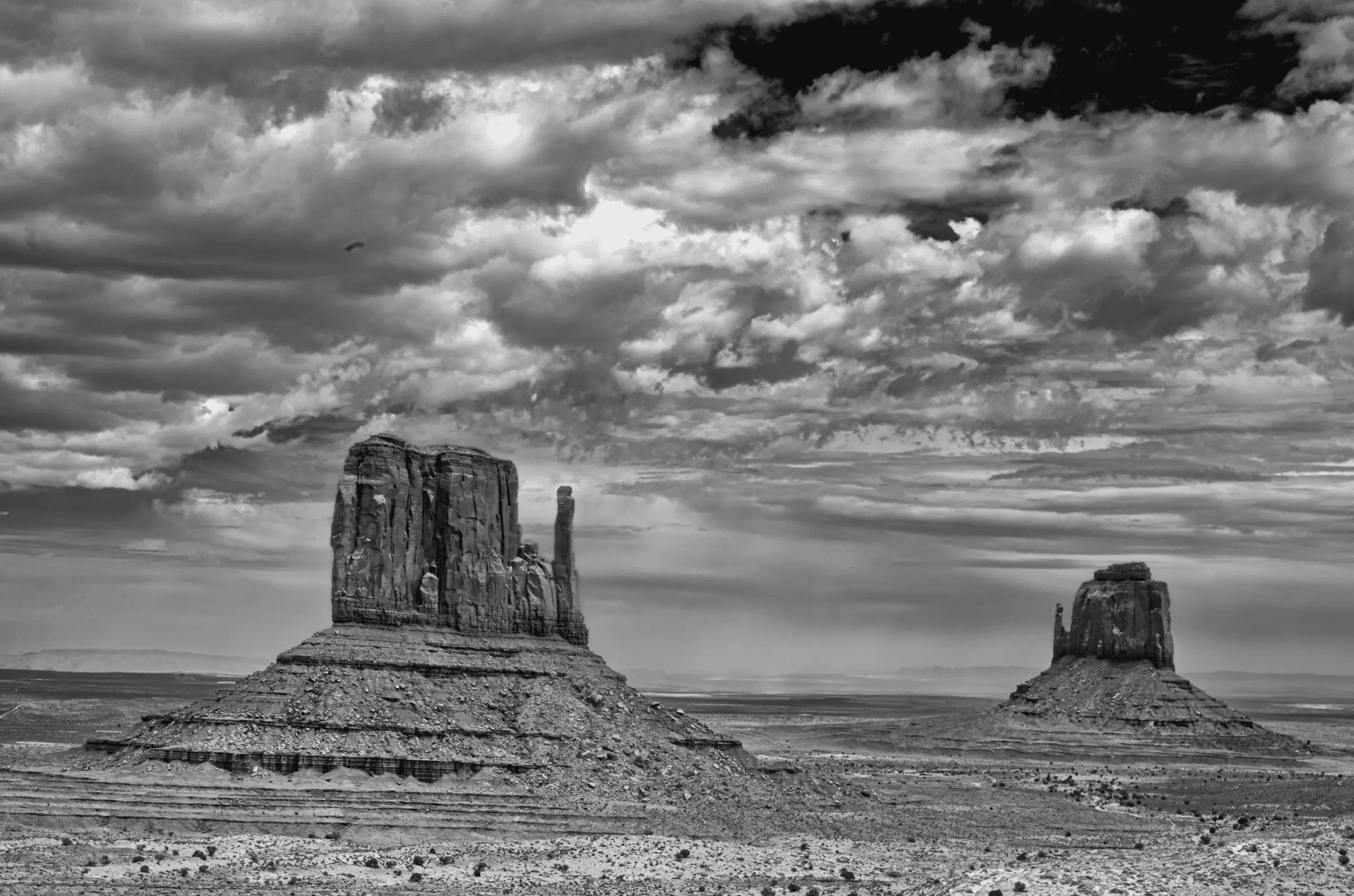 Two large sandstone buttes rise from a flat desert plain beneath a dramatic, cloud-filled sky.