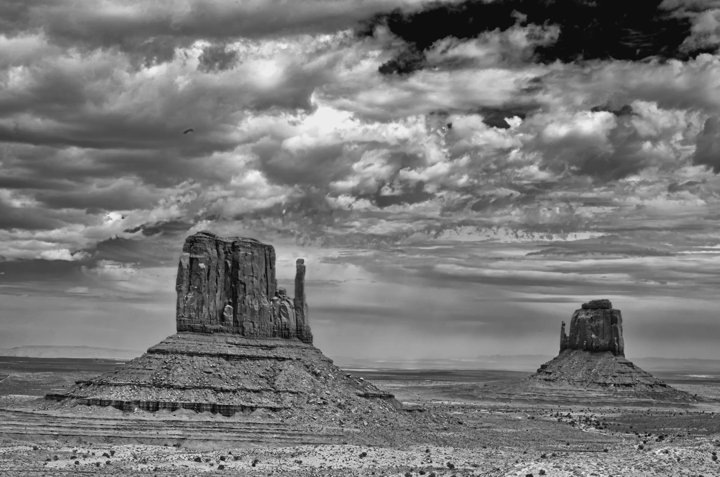 Click to view in fullscreen Two large sandstone buttes rise from a flat desert plain beneath a dramatic, cloud-filled sky.