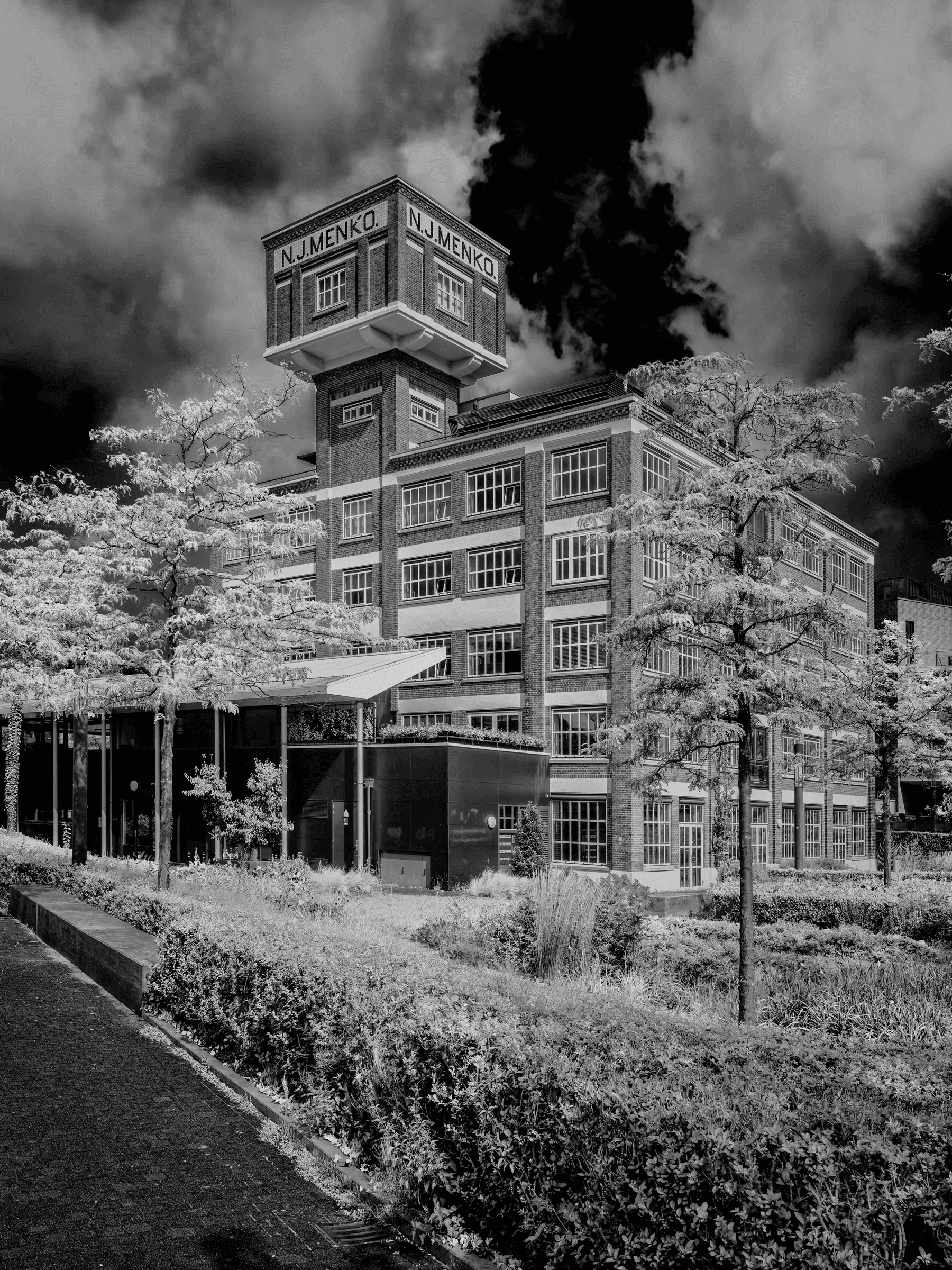 Click to view in fullscreen Black and white photograph of the N.J. Menko factory building in Enschede, with striking clouds and leafy trees in the foreground