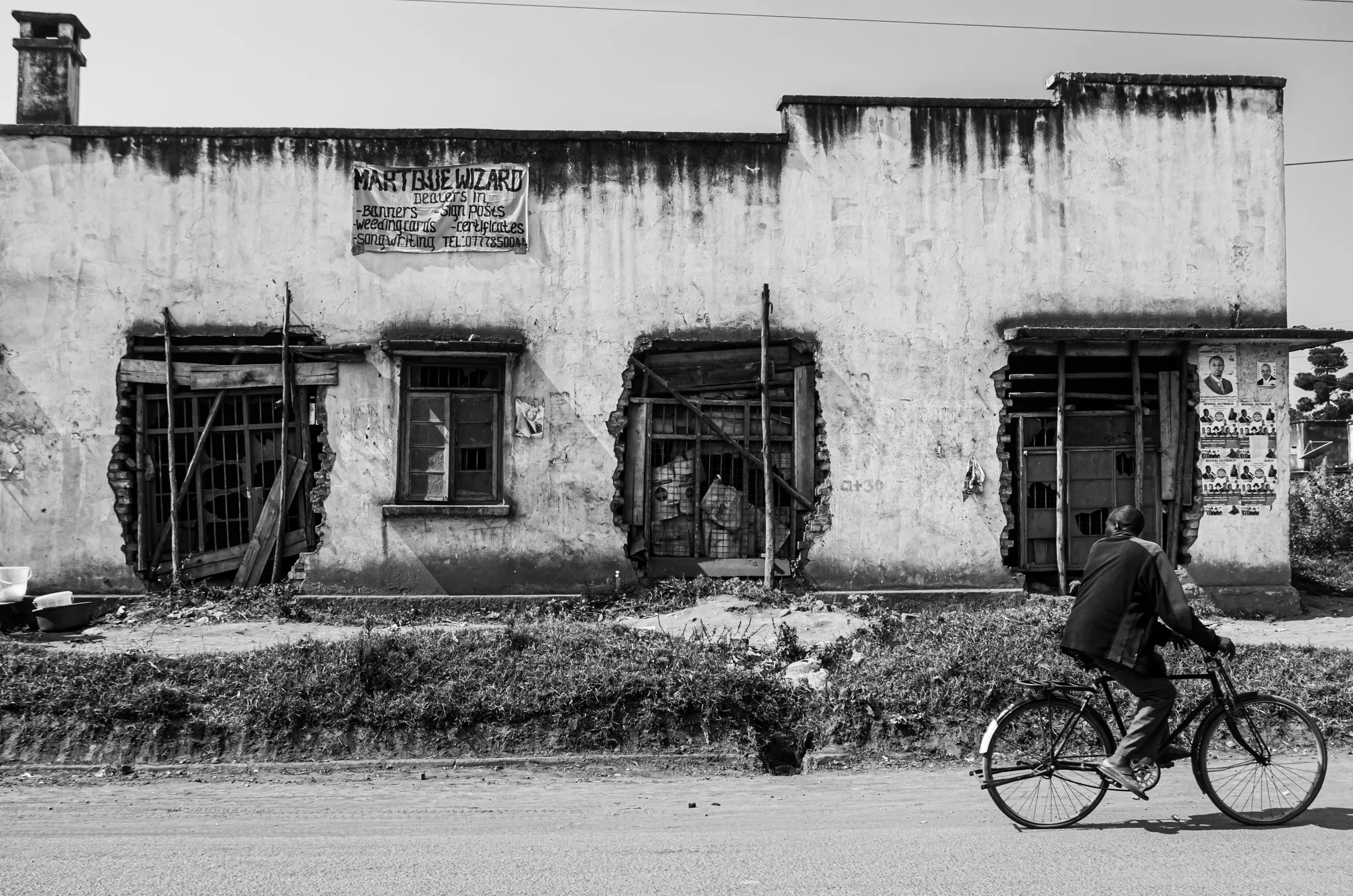 Black and white photograph of a person cycling past a weathered building with damaged windows on a roadside in Northern Uganda.