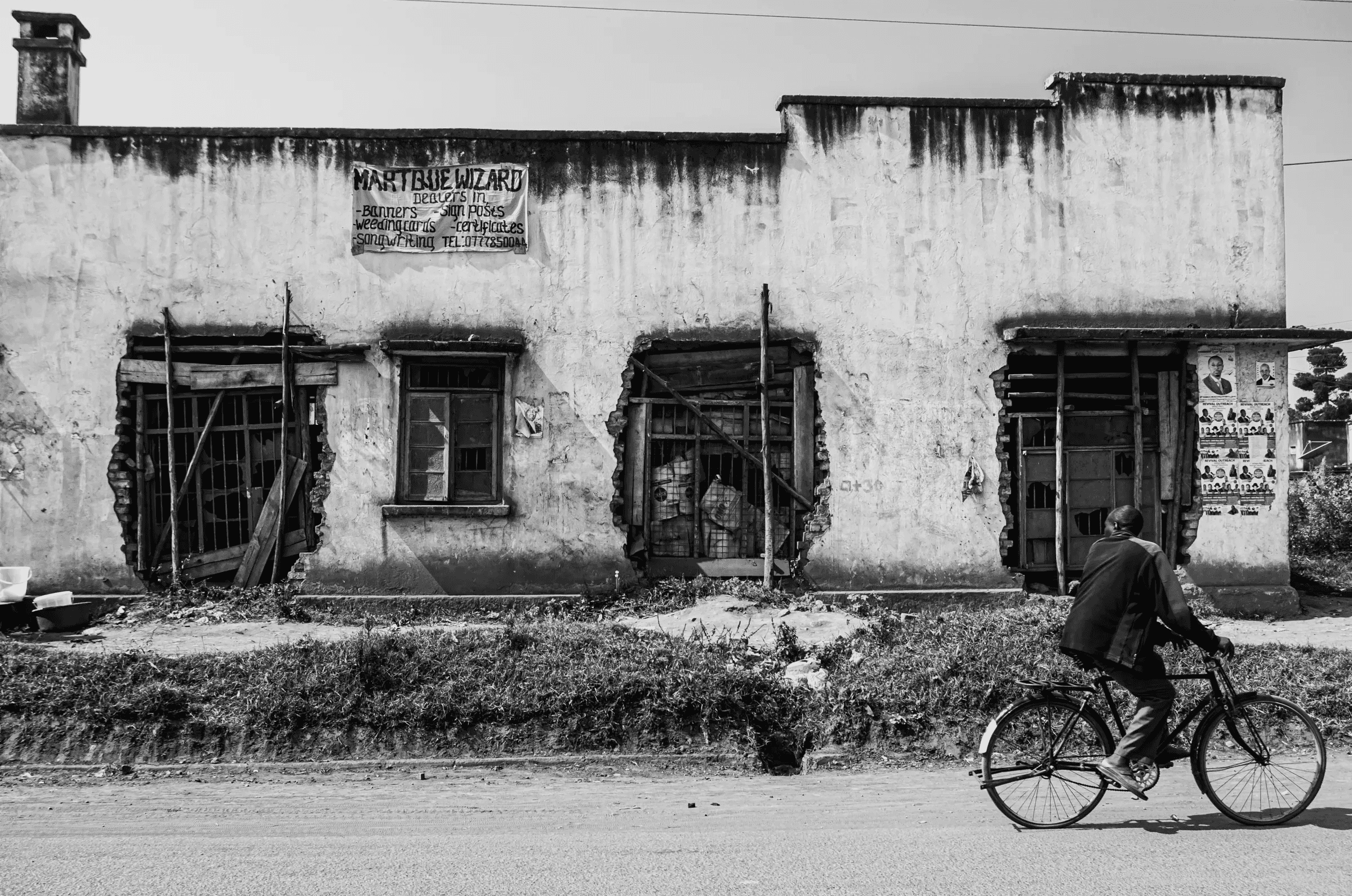 A person cycling past a weathered building with damaged windows on a roadside in Northern Uganda.