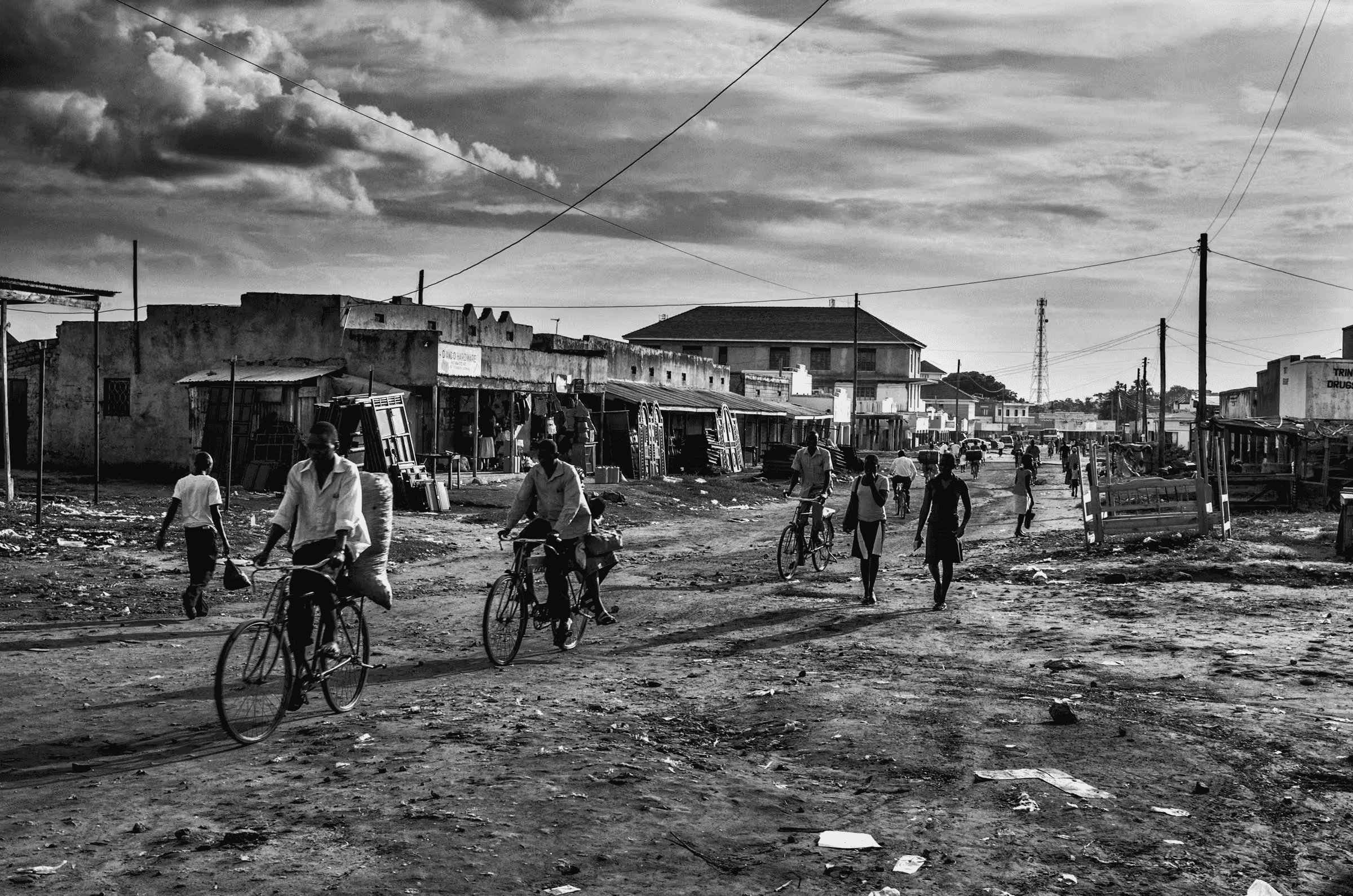 Black and white street scene photograph in a refugee settlement in Northern Uganda. People are walking and cycling on a dirt road lined with simple buildings and shops.