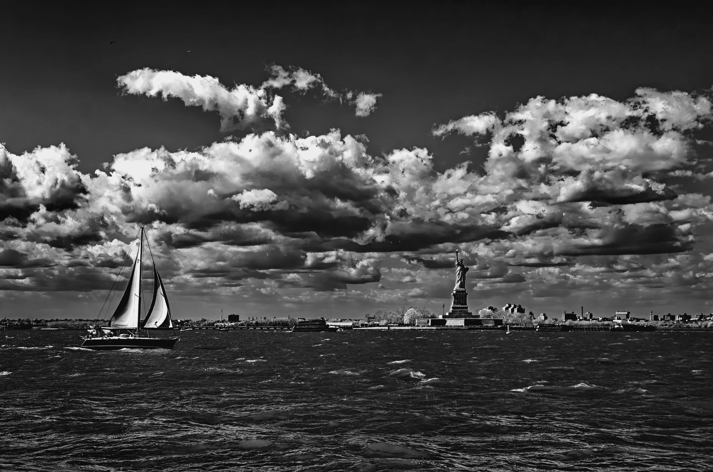 Dramatic black and white photograph of the Statue of Liberty and Lower Manhattan skyline viewed from New York Harbor, with a sailboat in the foreground under a cloudy sky.