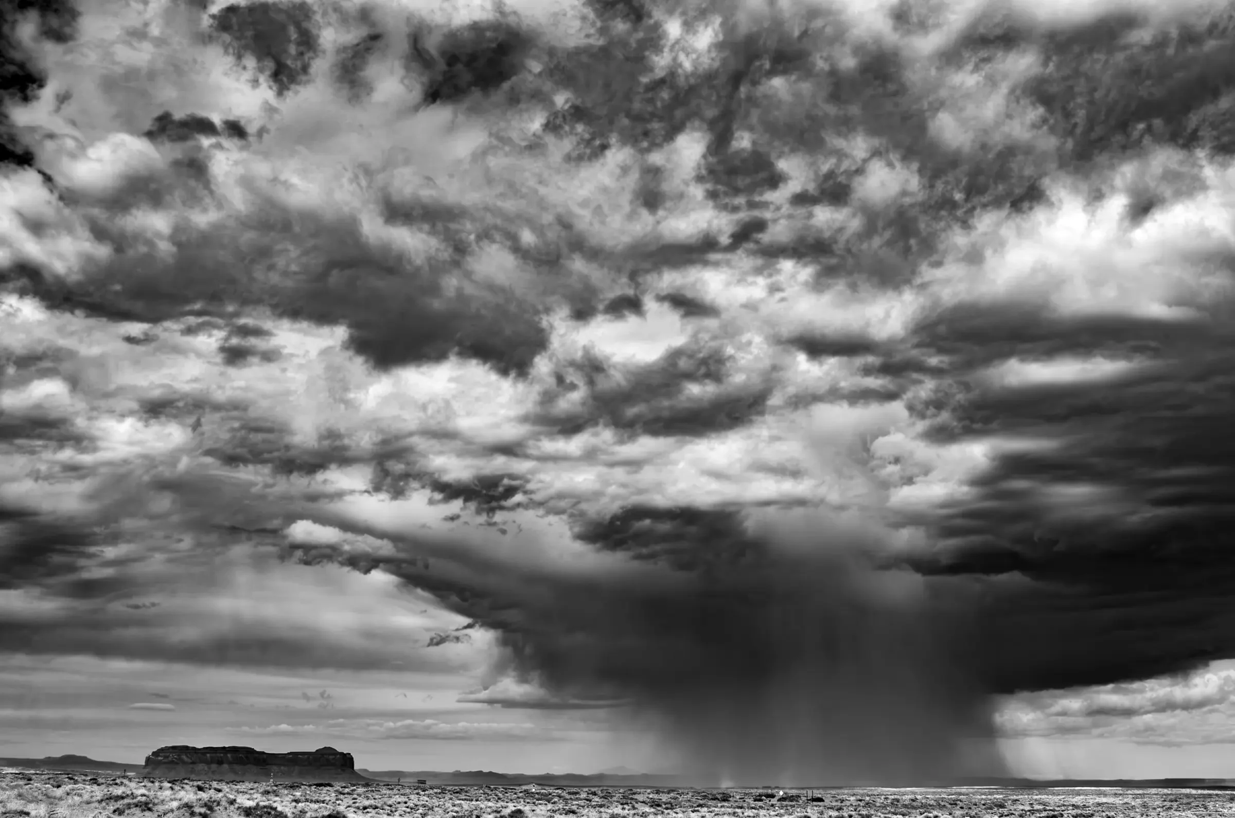 Black and white photograph of a dramatic sky over a flat desert landscape, with a heavy rain shaft falling from dark clouds near a distant mesa.