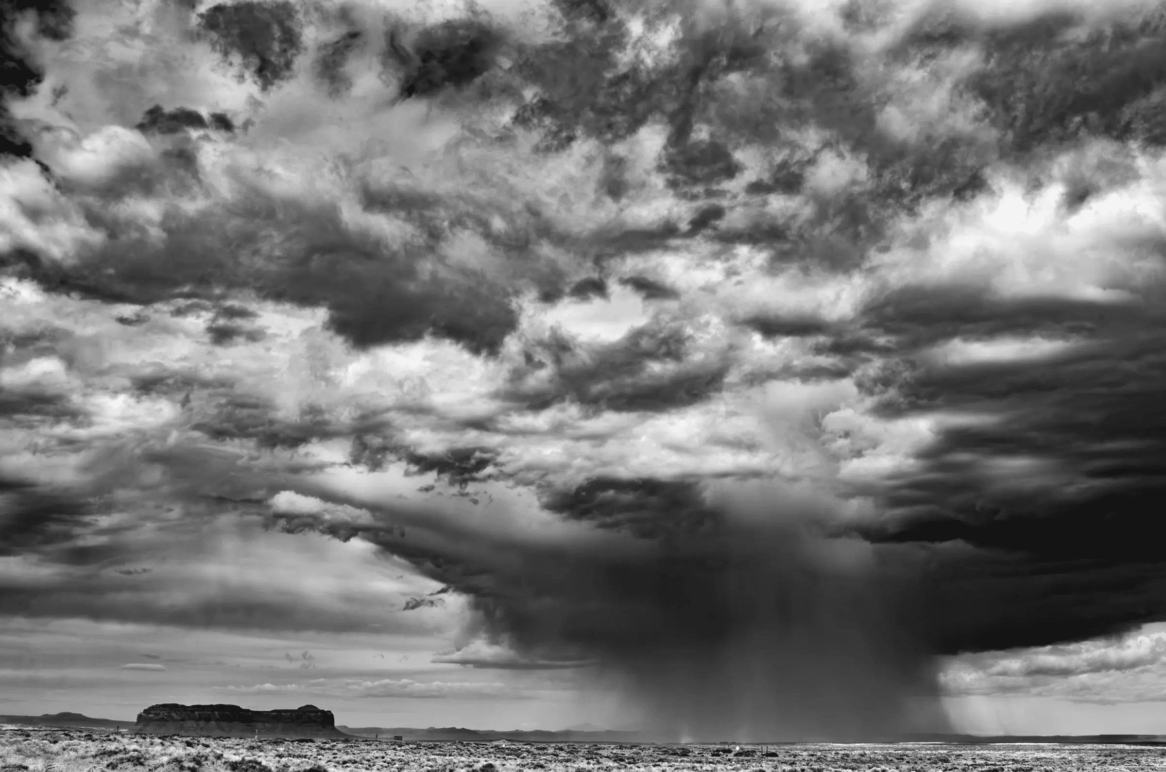 Black and white photograph of a dramatic sky over a flat desert landscape, with a heavy rain shaft falling from dark clouds near a distant mesa.