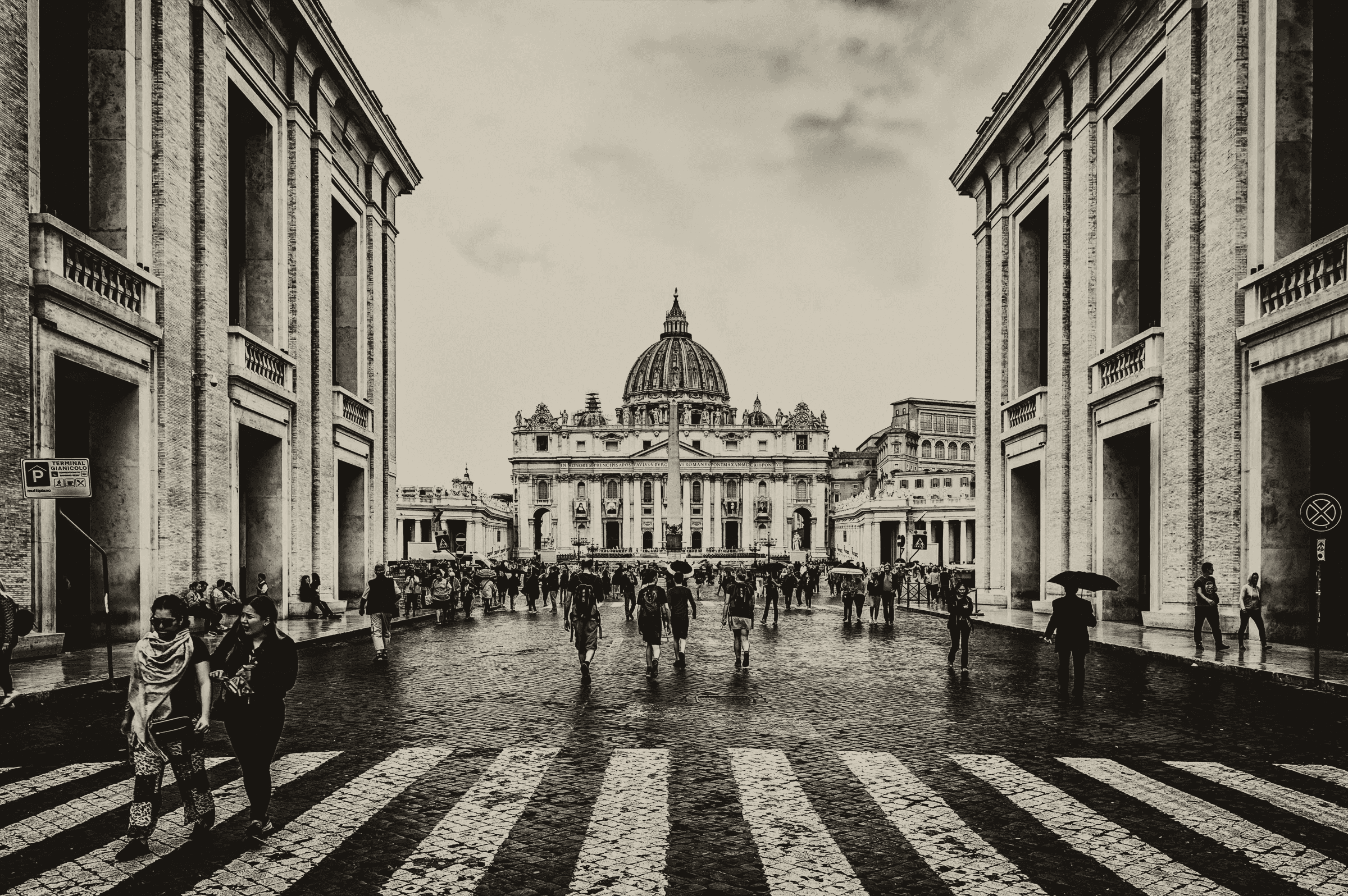 Monochrome photograph looking down the Via della Conciliazione towards St. Peter's Basilica and Square in Vatican City, with numerous pedestrians walking on the cobblestone street.