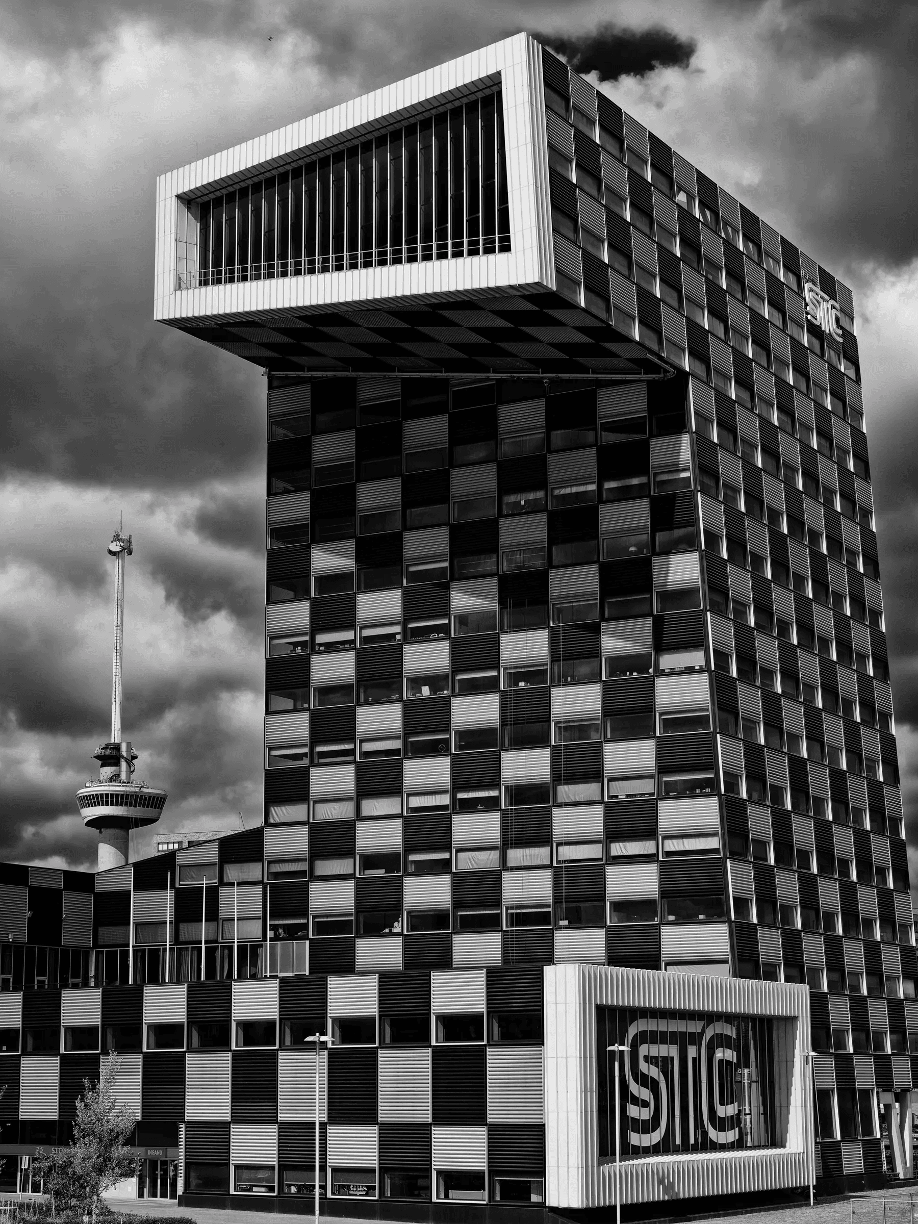 Click to view in fullscreen Black and white photograph of the STC building in Rotterdam with its bold geometric design, set against a moody sky and the Euromast tower in the background.