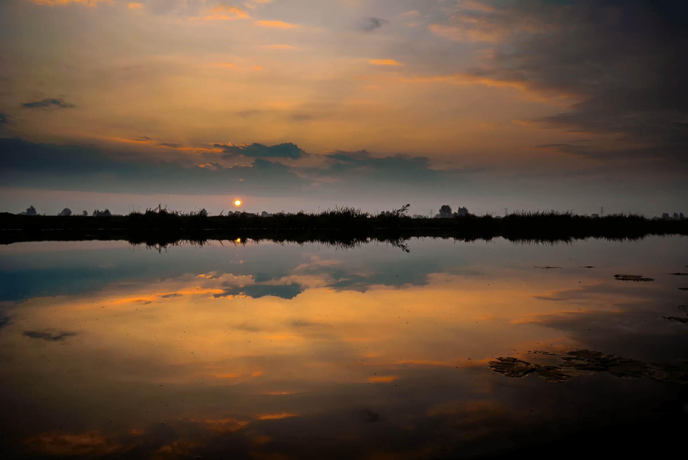 Click to view in fullscreen Vibrant sunset sky with orange and dark clouds reflected in calm water.