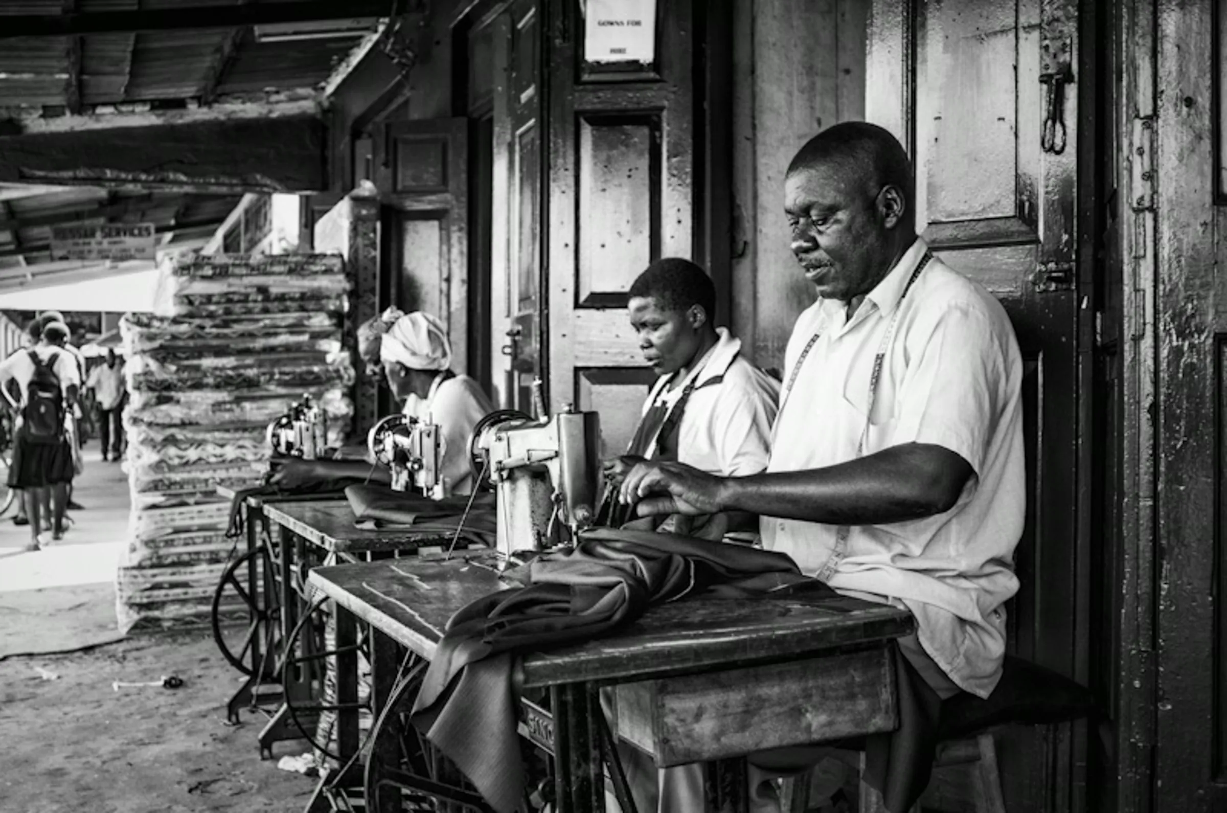 Black and white photograph of three people, two women and a man, working intently at sewing machines set up outdoors, possibly in a market or workshop.