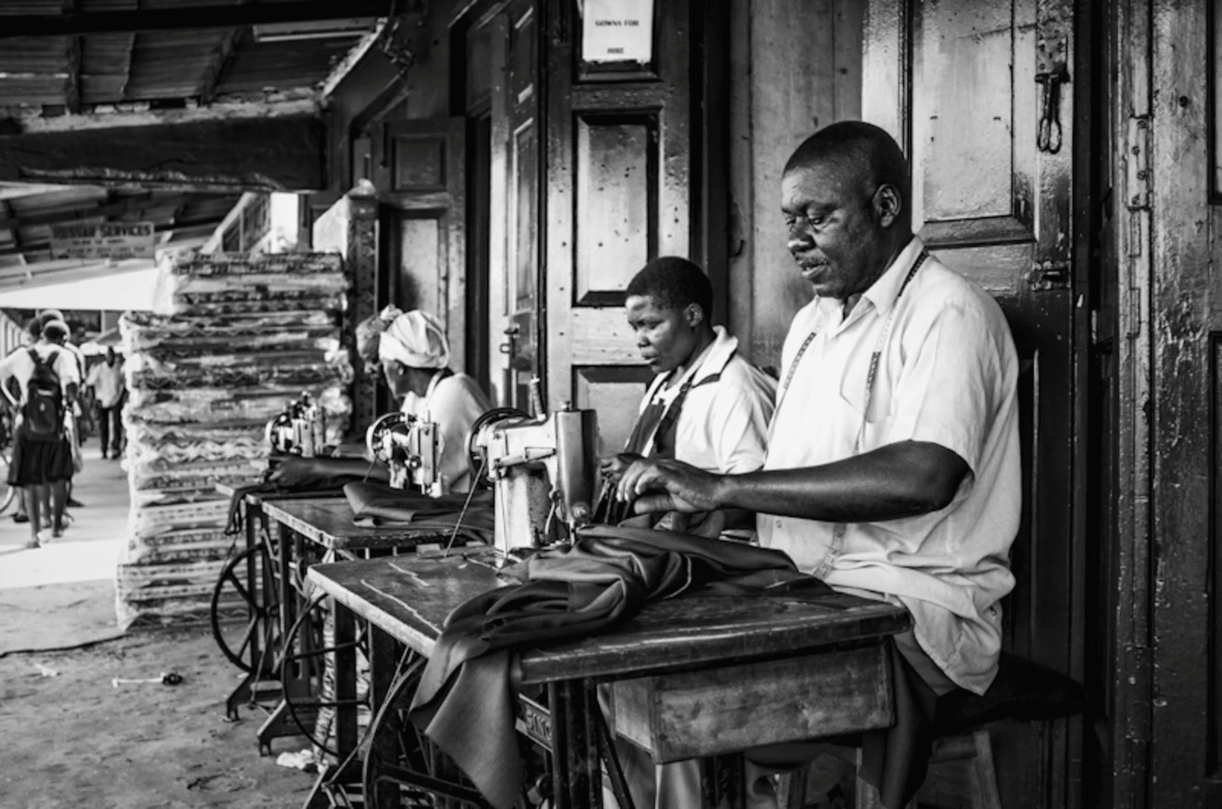 Black and white photograph of three people, two women and a man, working intently at sewing machines set up outdoors, possibly in a market or workshop.