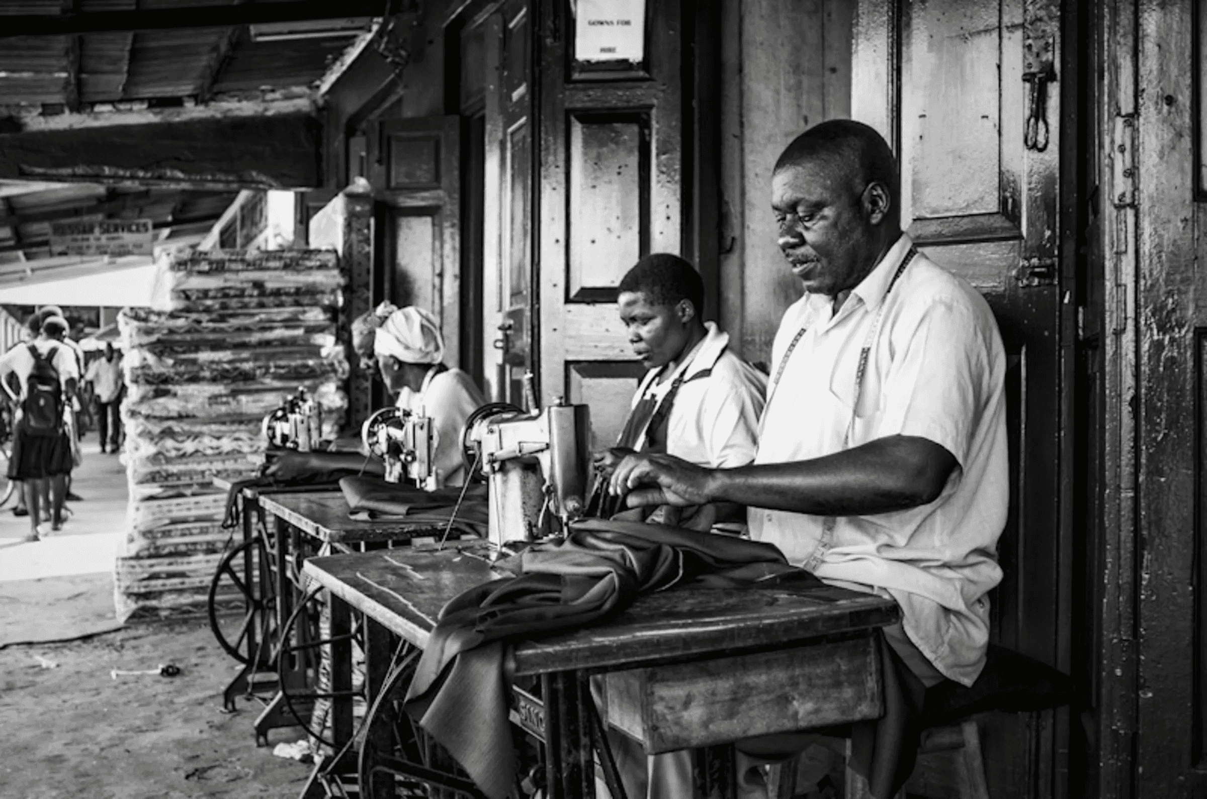 Black and white photograph of three people, two women and a man, working intently at sewing machines set up outdoors, possibly in a market or workshop.