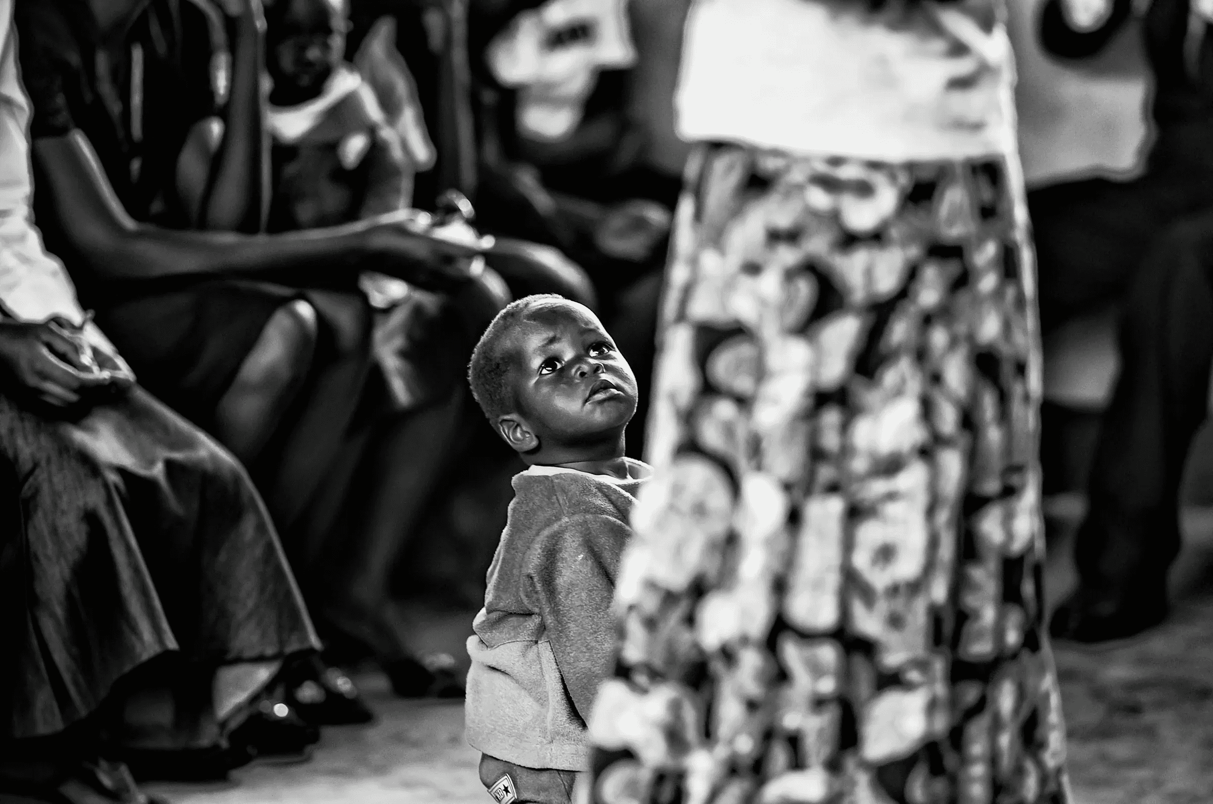 Black and white photograph focusing on a small Ugandan child looking upwards, partially obscured by an adult figure in the foreground.