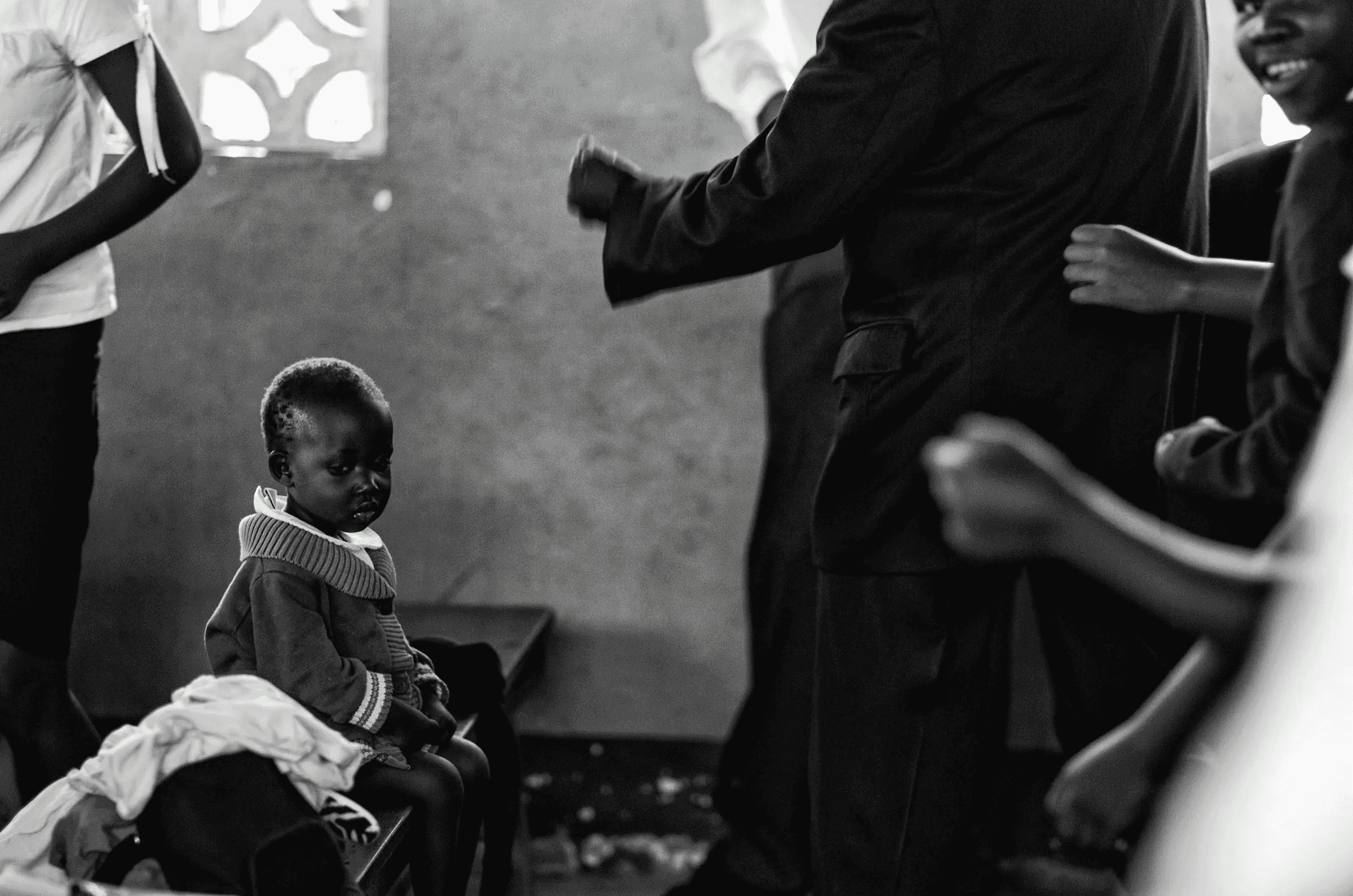 Black and white photograph of a small Ugandan child sitting quietly on a bench, observing activity in the background where other people are gathered indoors.