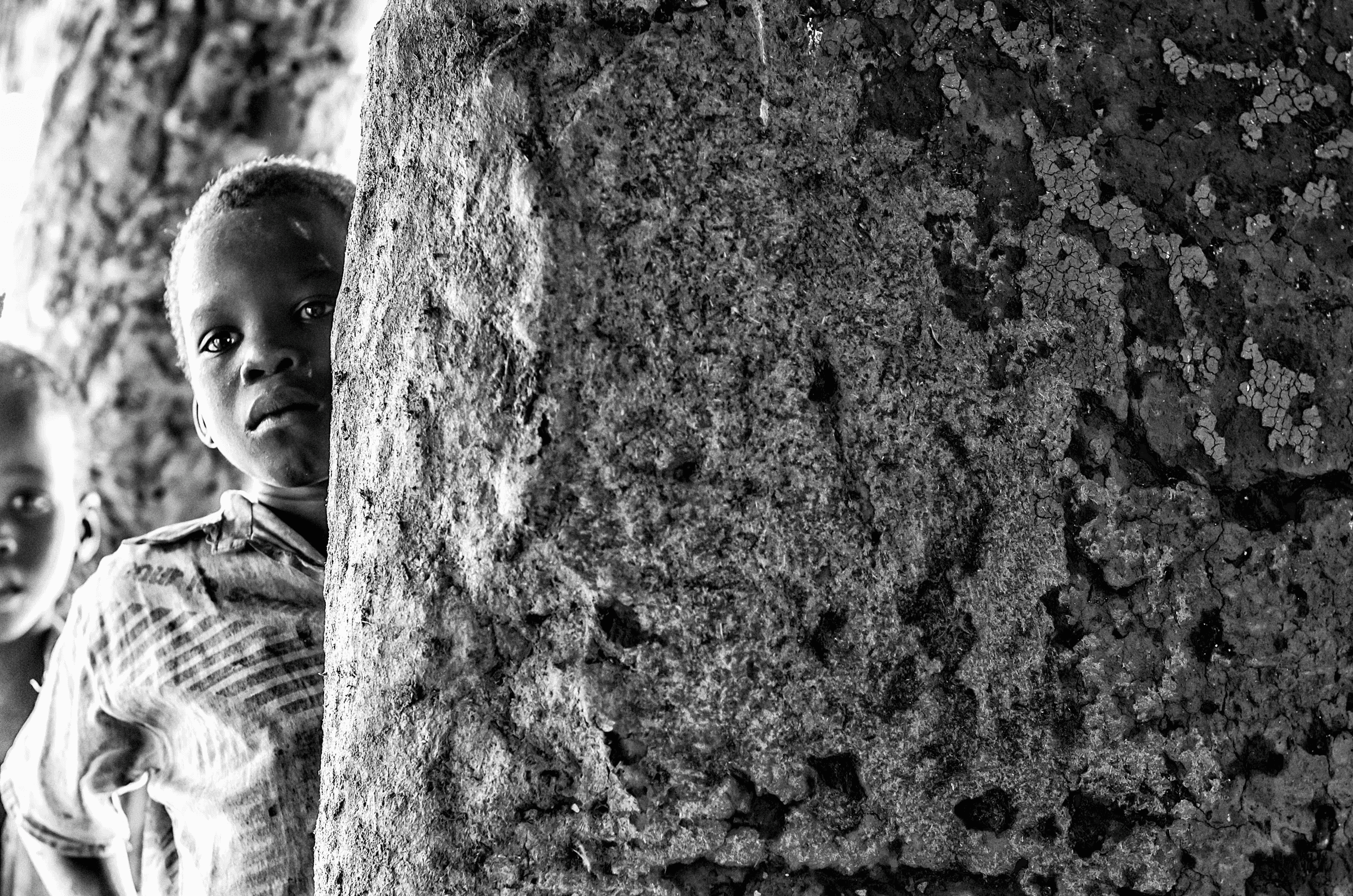 Click to view in fullscreen A young Ugandan child peeking around a rough, textured pillar or wall, looking towards the camera.