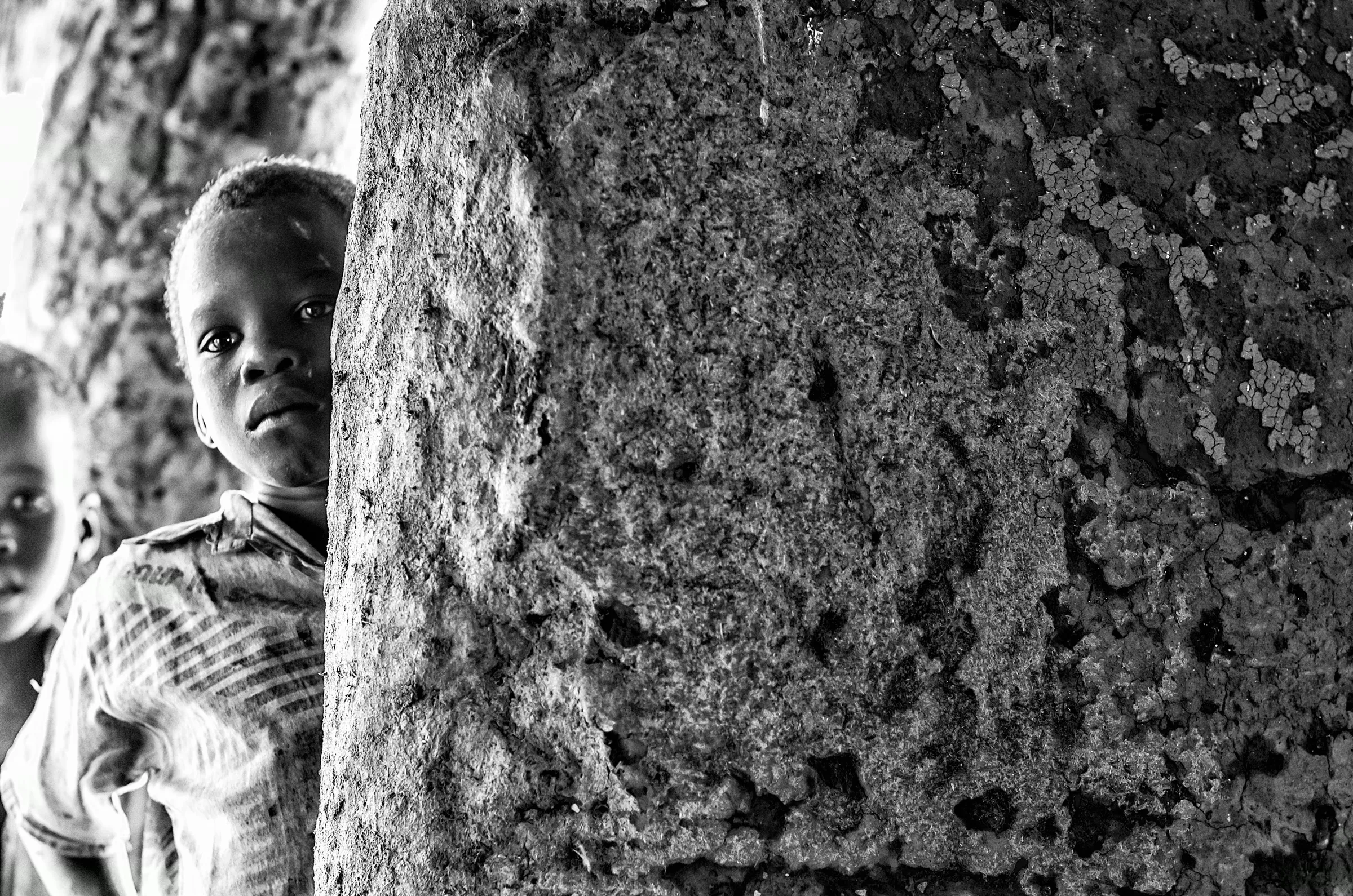 A young Ugandan child peeking around a rough, textured pillar or wall, looking towards the camera.