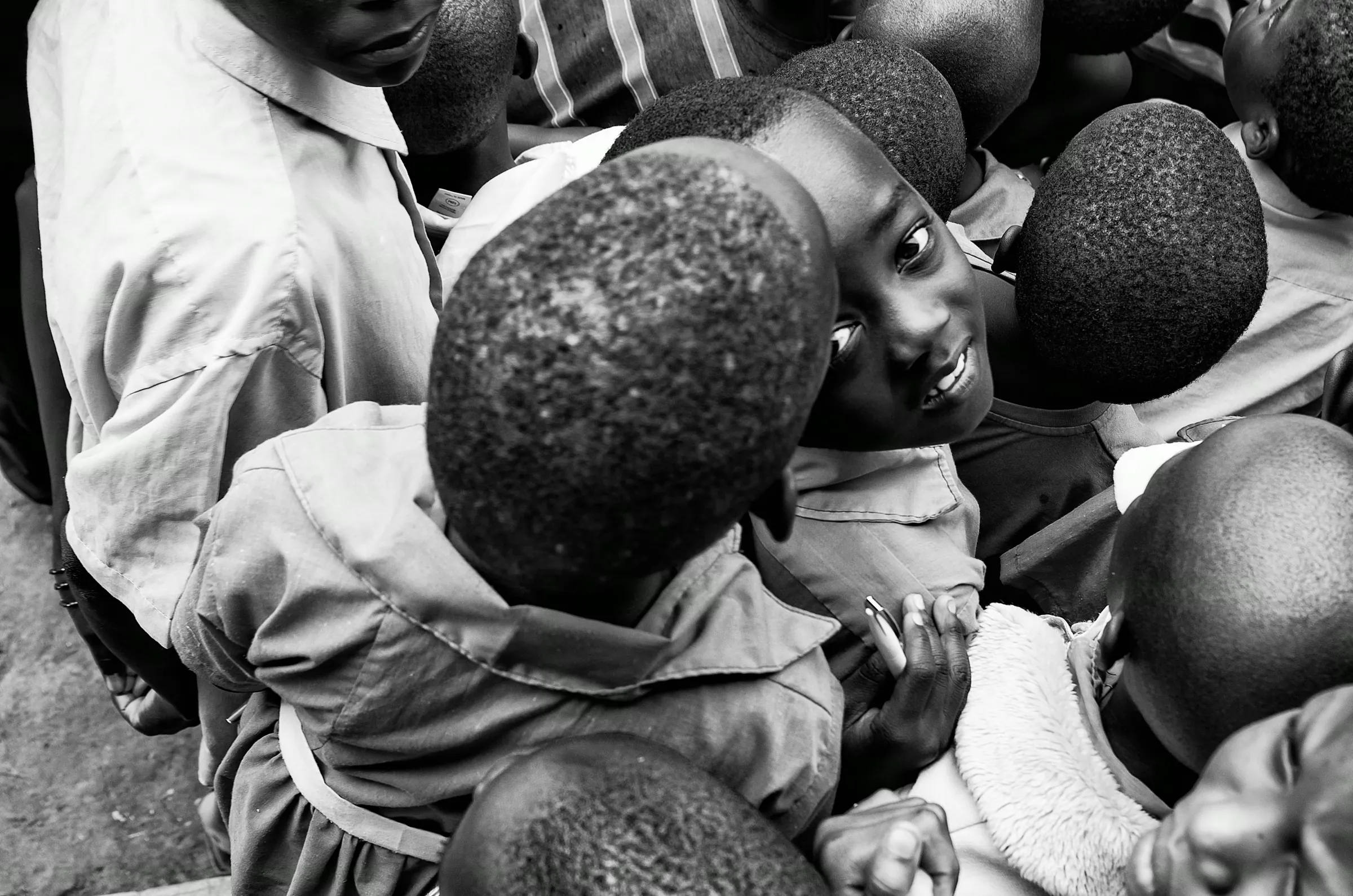 Black and white photograph taken from a high angle, looking down into a group of Ugandan children, one of whom looks directly up at the camera.