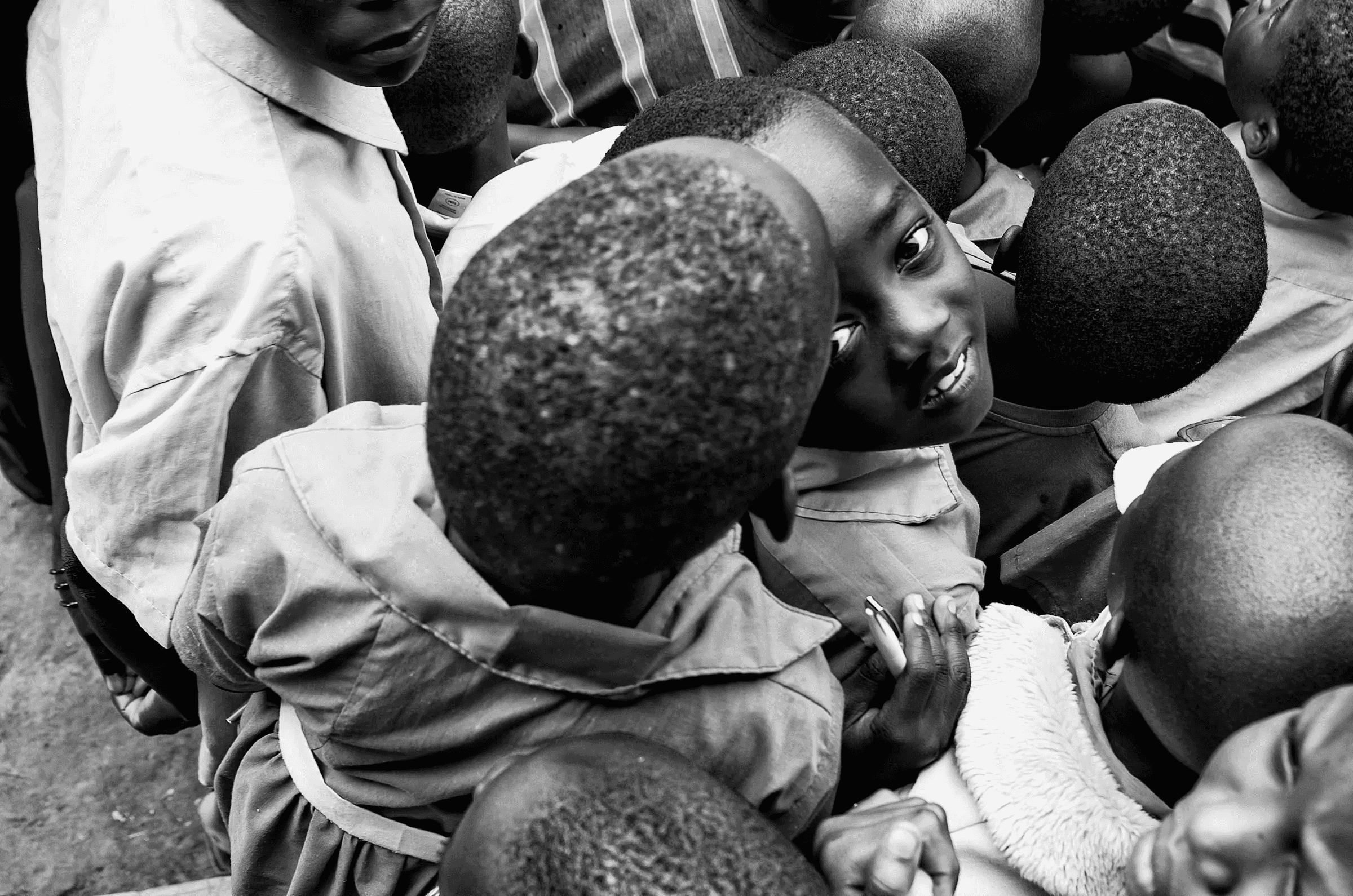 Black and white photograph taken from a high angle, looking down into a group of Ugandan children, one of whom looks directly up at the camera.
