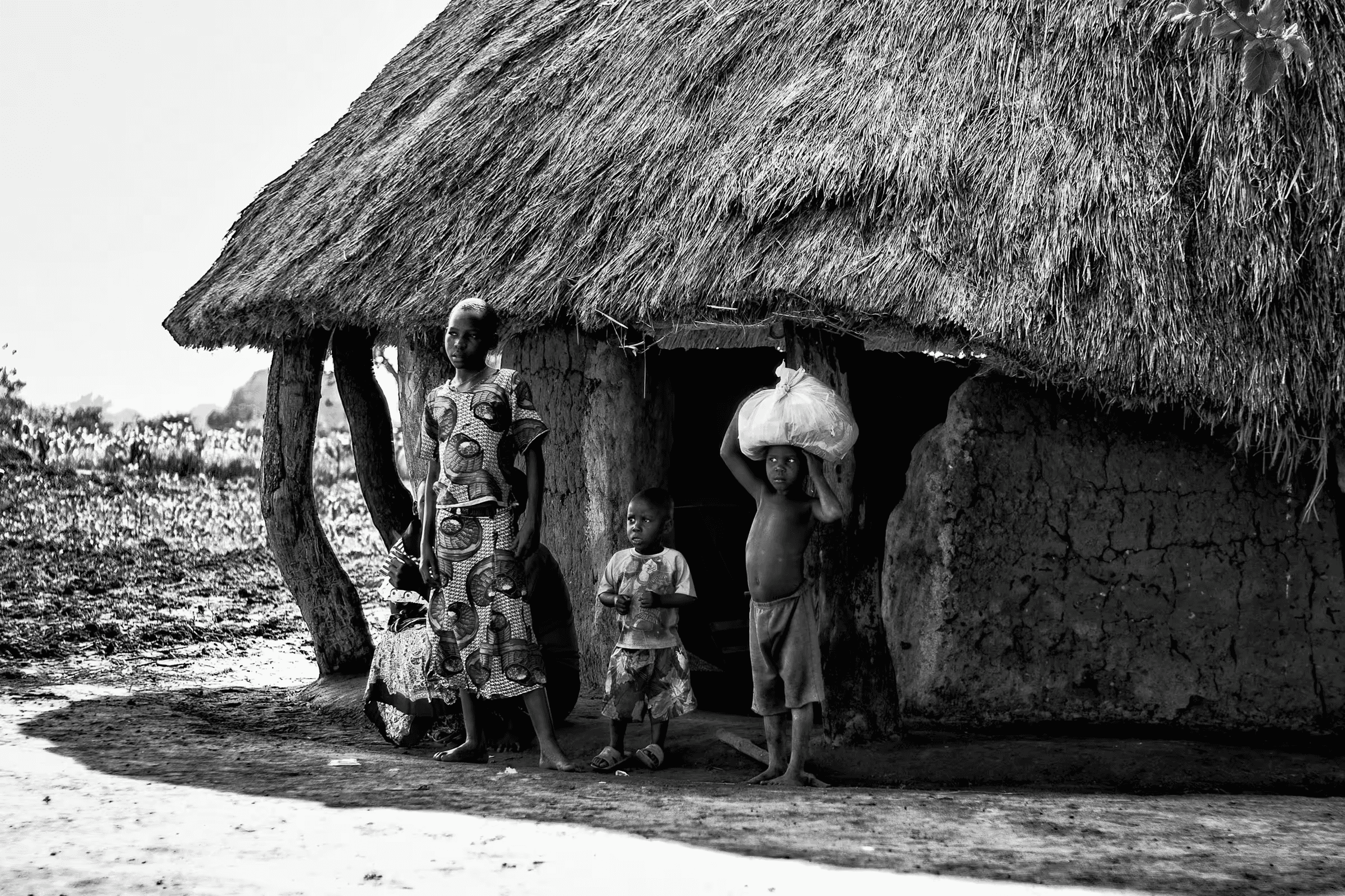 Black and white photograph of a Ugandan family standing outside their traditional mud hut with a thatched roof.