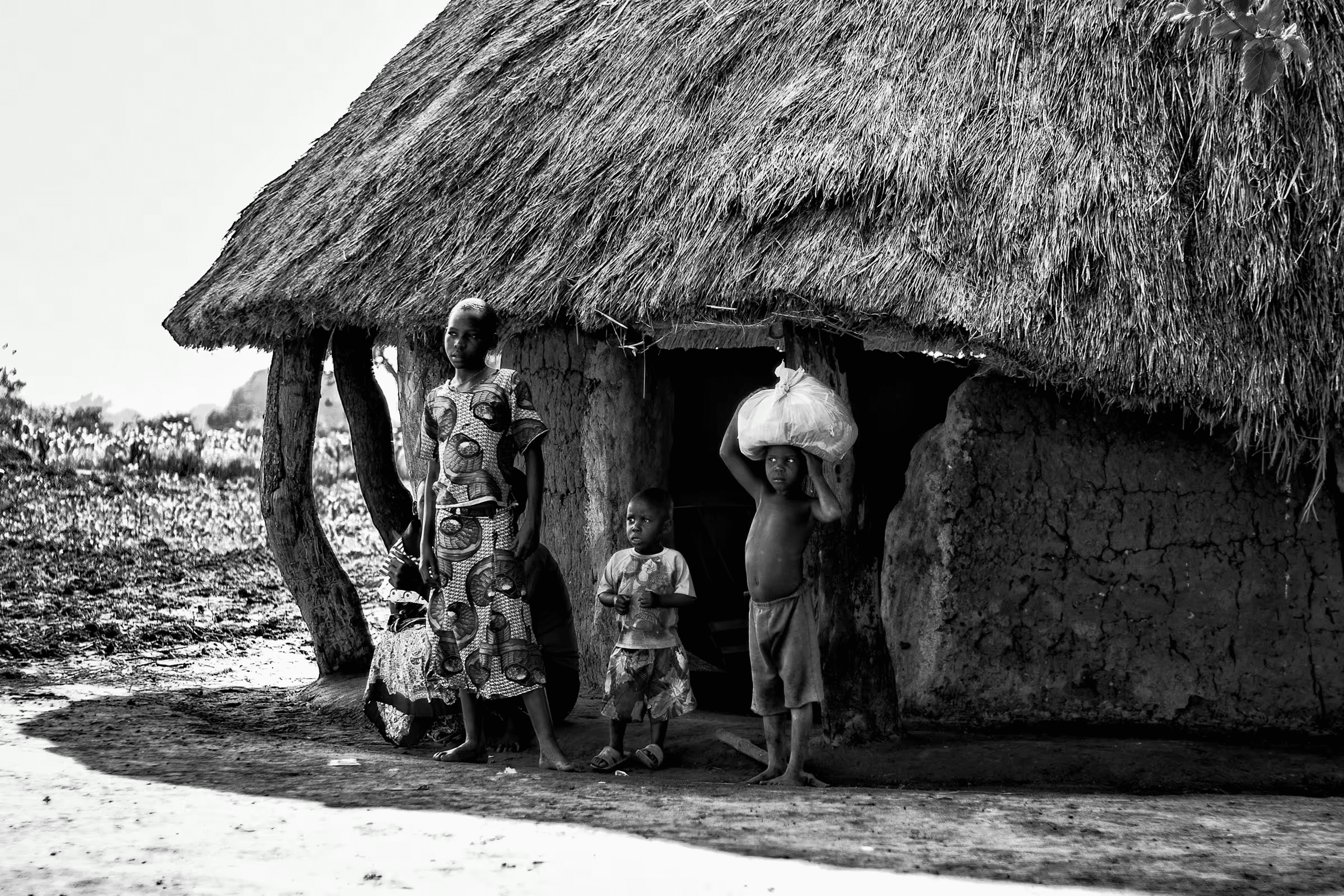 Black and white photograph of a Ugandan family standing outside their traditional mud hut with a thatched roof.
